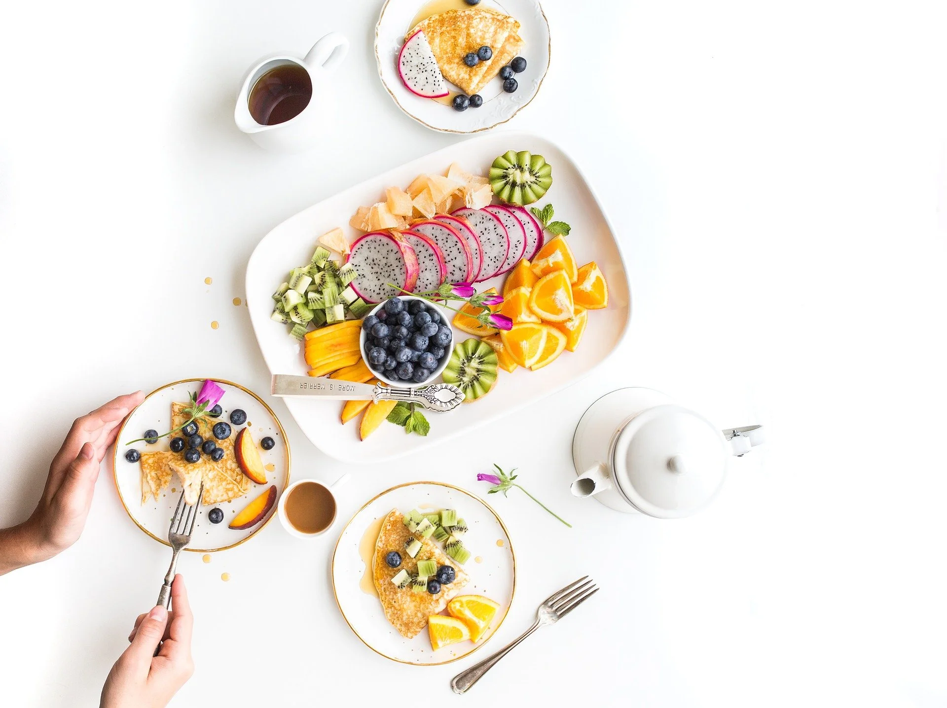 A breakfast setup with a large white platter of sliced fruits including dragon fruit, blueberries, kiwi, oranges, and watermelon, along with a small bowl of blueberries. Two small plates hold pancakes topped with blueberries, kiwi, and orange slices. There is a white teapot, a small white pitcher of syrup, two forks, and the hand of a person reaching for one of the pancakes.