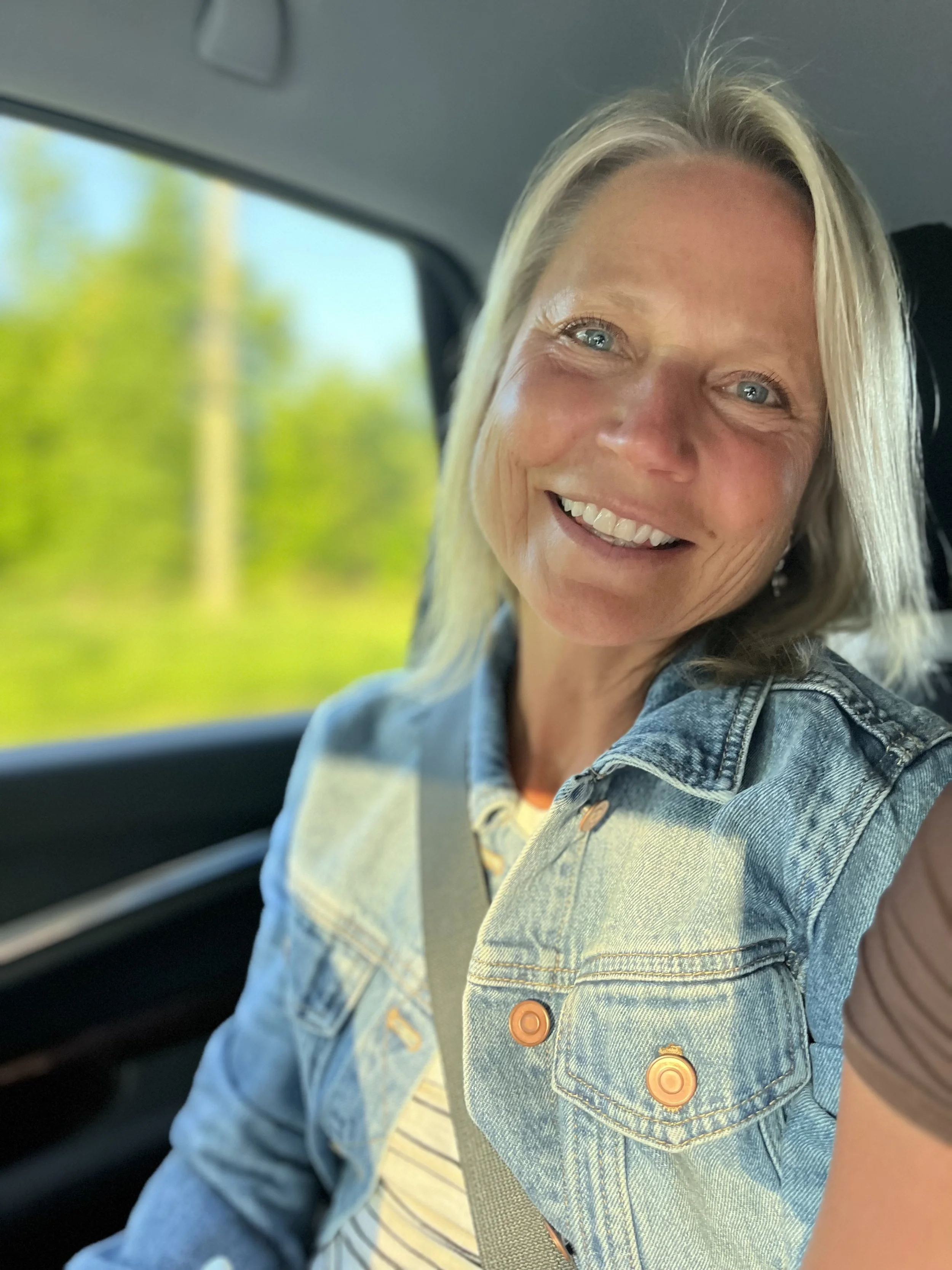 A smiling woman with blonde hair and blue eyes wearing a denim jacket, sitting in a car with a seatbelt on, with green trees visible through the window.