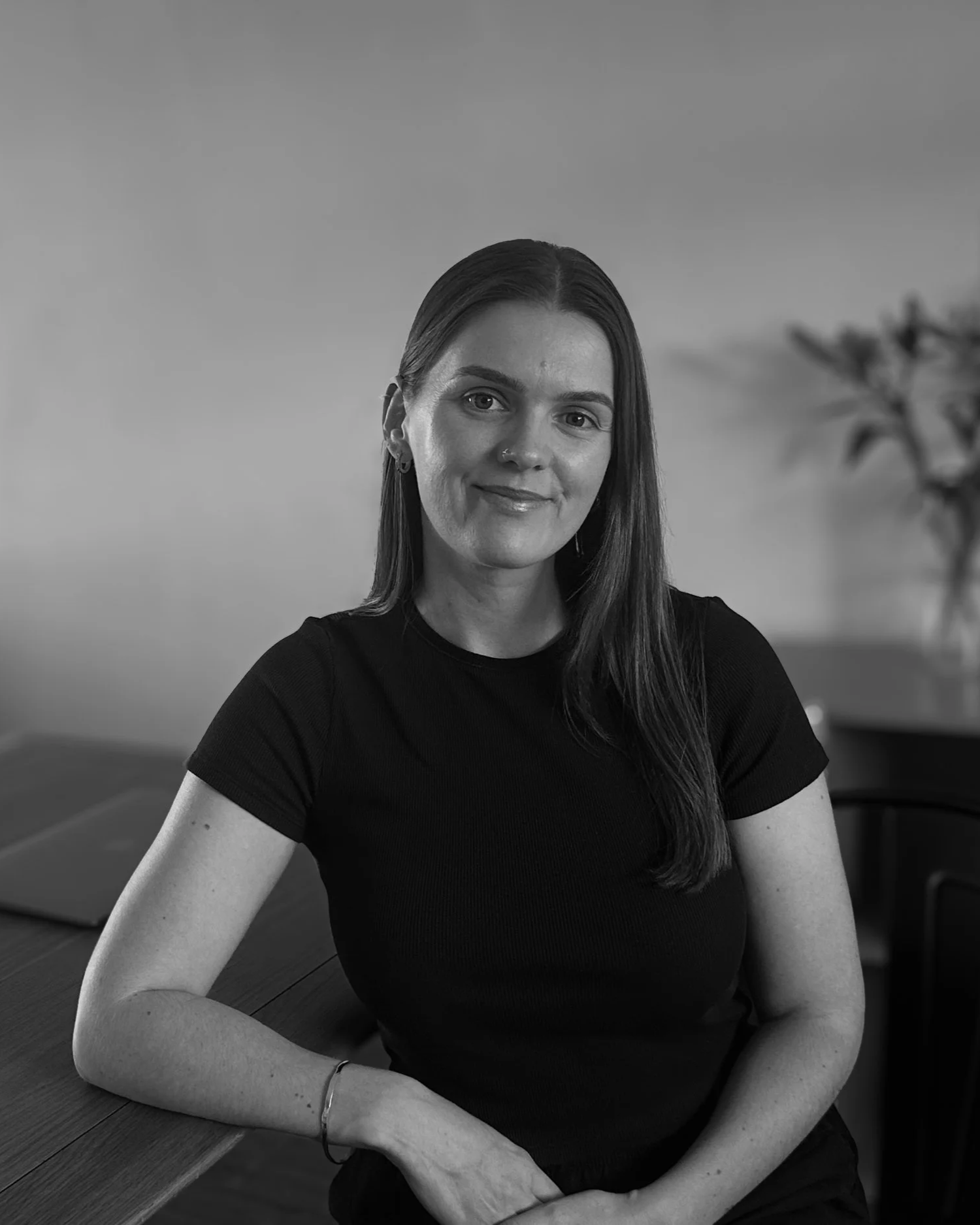A woman with straight dark hair appears to be sitting at a table, wearing a black short-sleeved shirt, and smiling softly in a black-and-white photo.