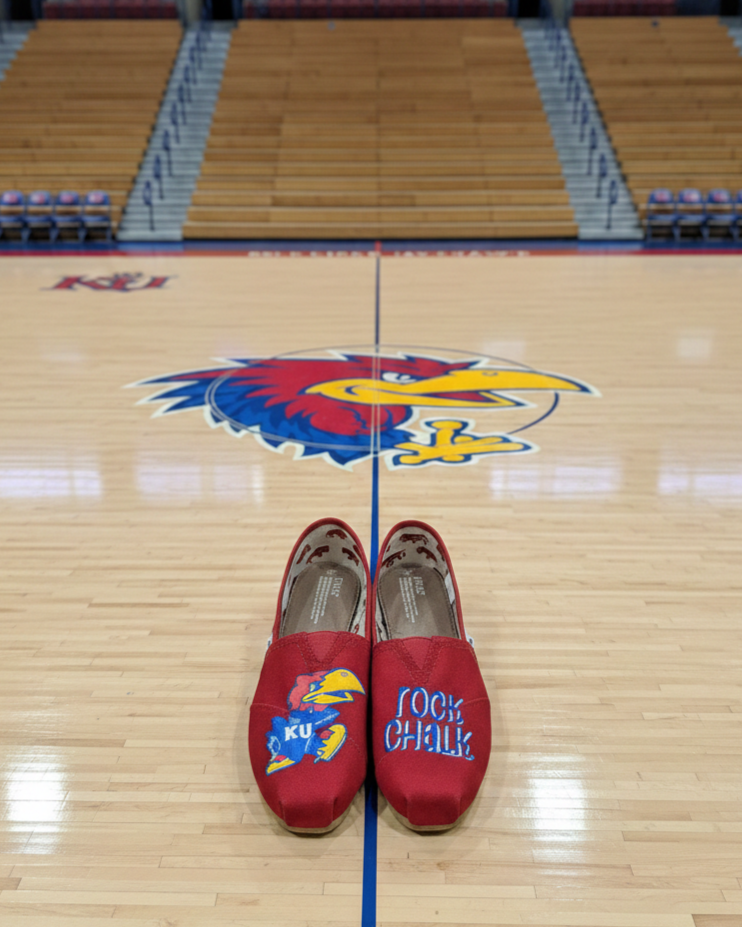 A pair of red slippers with Kansas Jayhawks logos and the words "Rock Chalk" on the toes, placed on a basketball court with the Jayhawks logo at the center.