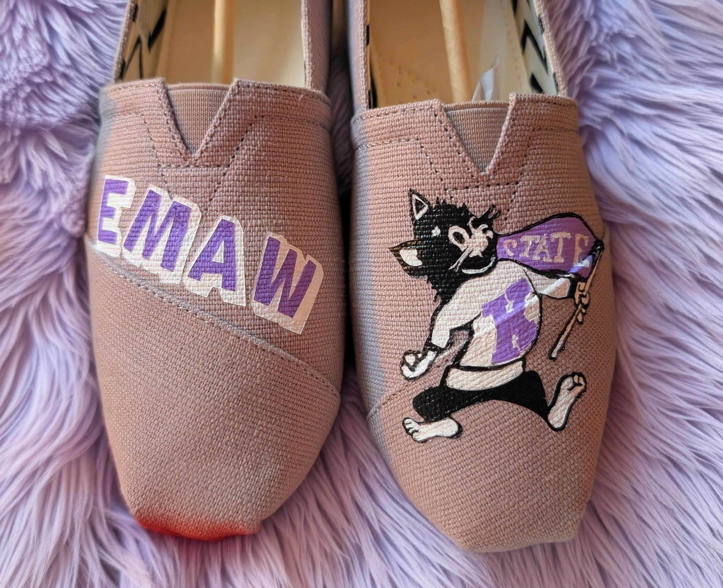 Pink slip-on sneakers with a cartoon of a black and white cat holding a police baton, and the words 'EMAW' and 'STATE' on them, placed on a purple furry surface.