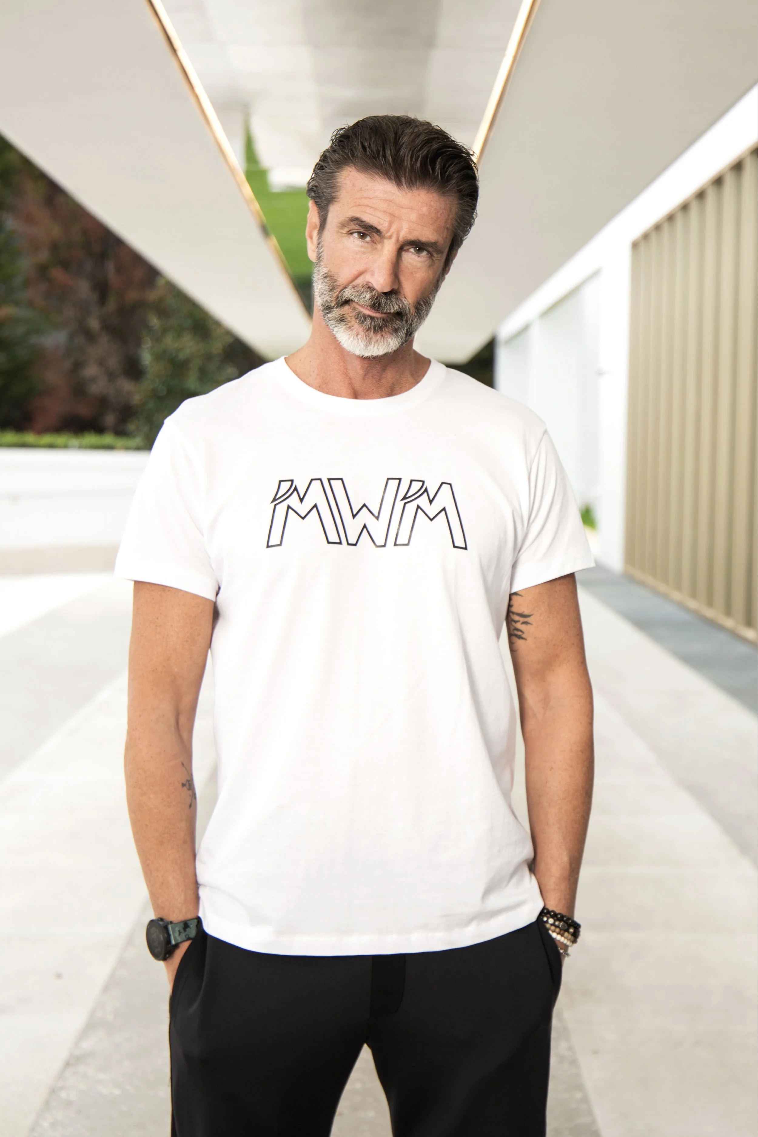 Uomo con barba e capelli grigi, indossa una maglietta bianca con scritta MKWM, posa con le mani nelle tasche davanti a un edificio moderno con linee geometriche e luci.