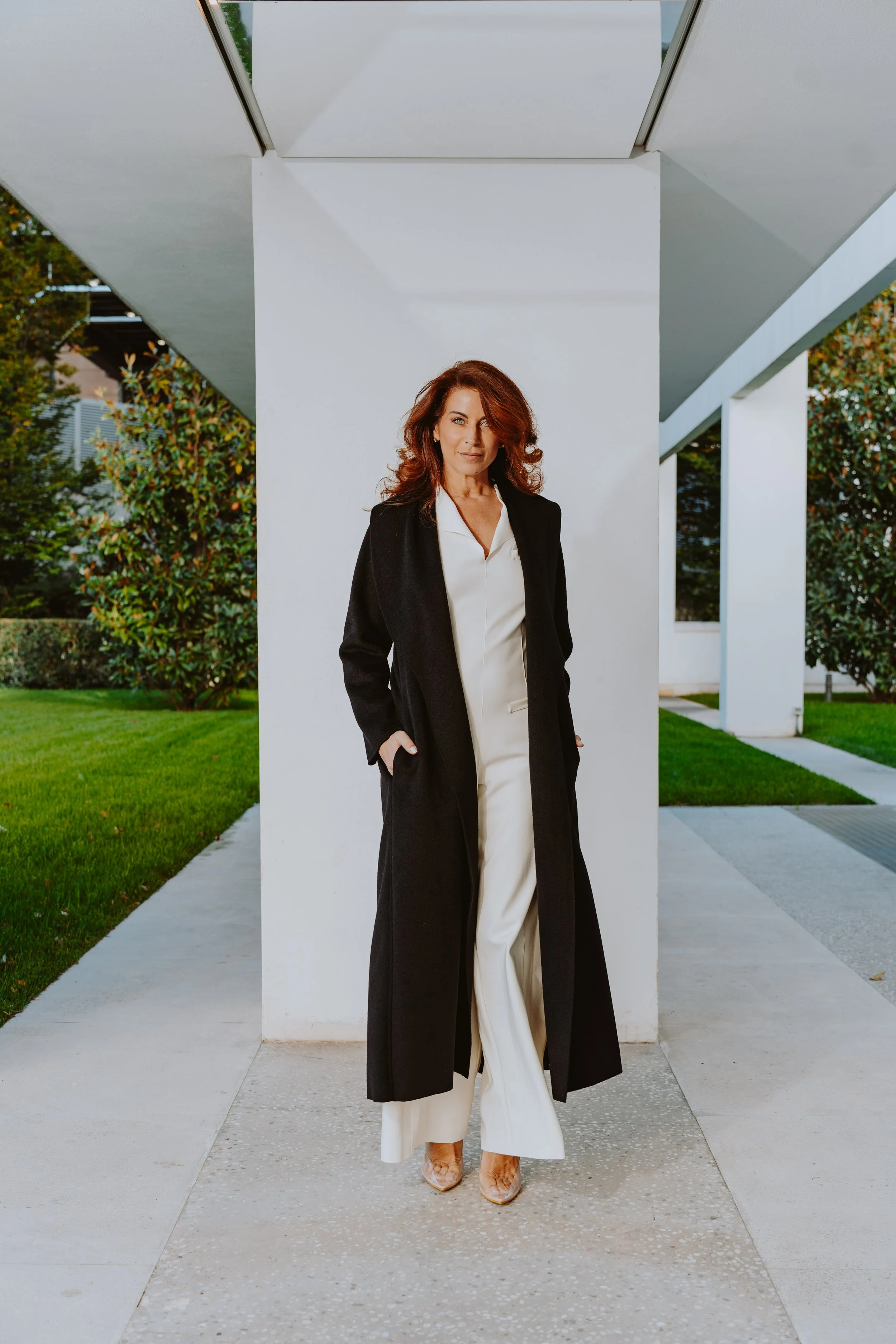 Donna con capelli ricci in piedi davanti a una parete bianca all'aperto, indossa un completo bianco e un cappotto nero, con scarpe a tacco.
