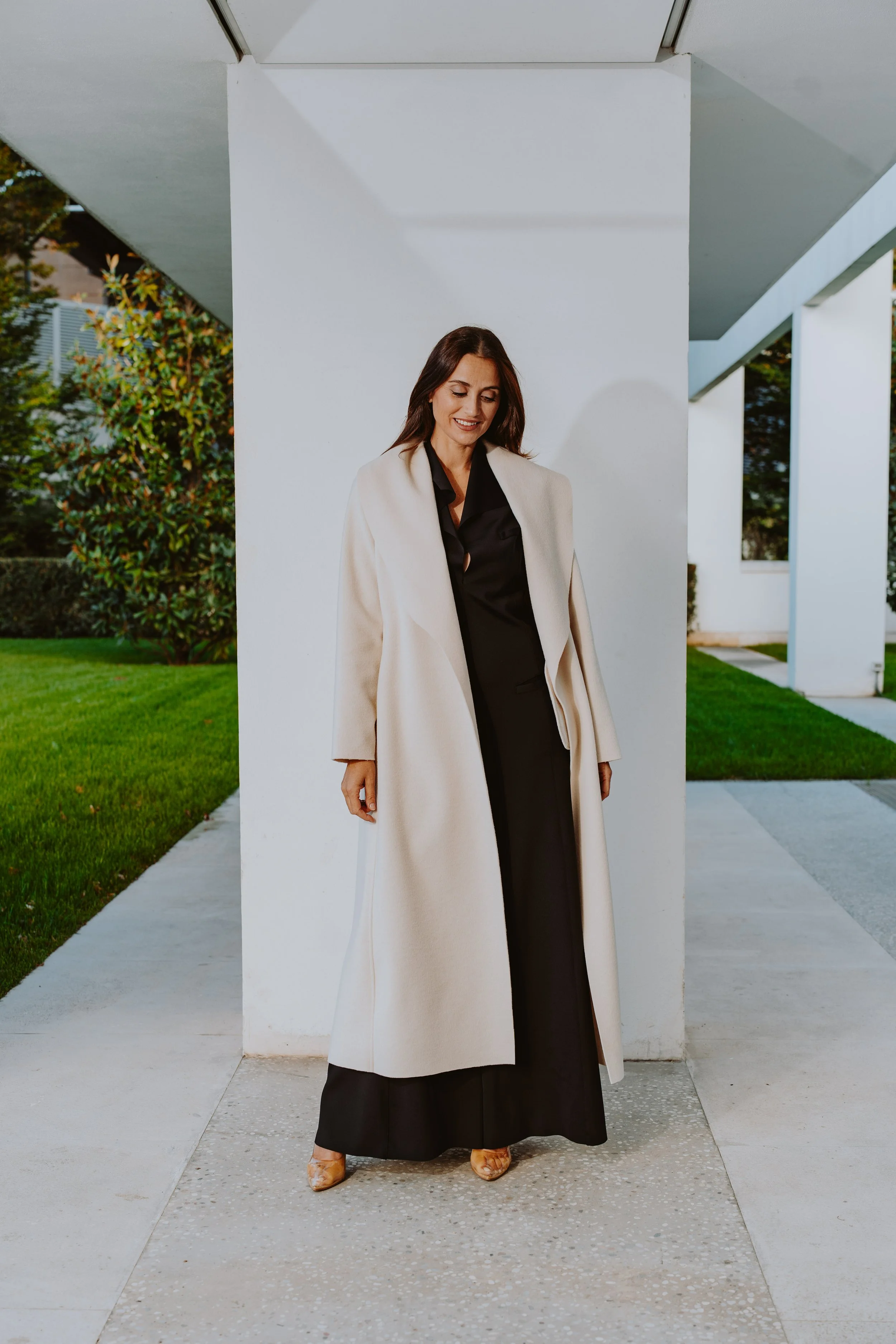 Una donna elegante con un cappotto crema e un vestito nero, che si trova all'aperto davanti a una struttura moderna bianca con un giardino verde sullo sfondo.