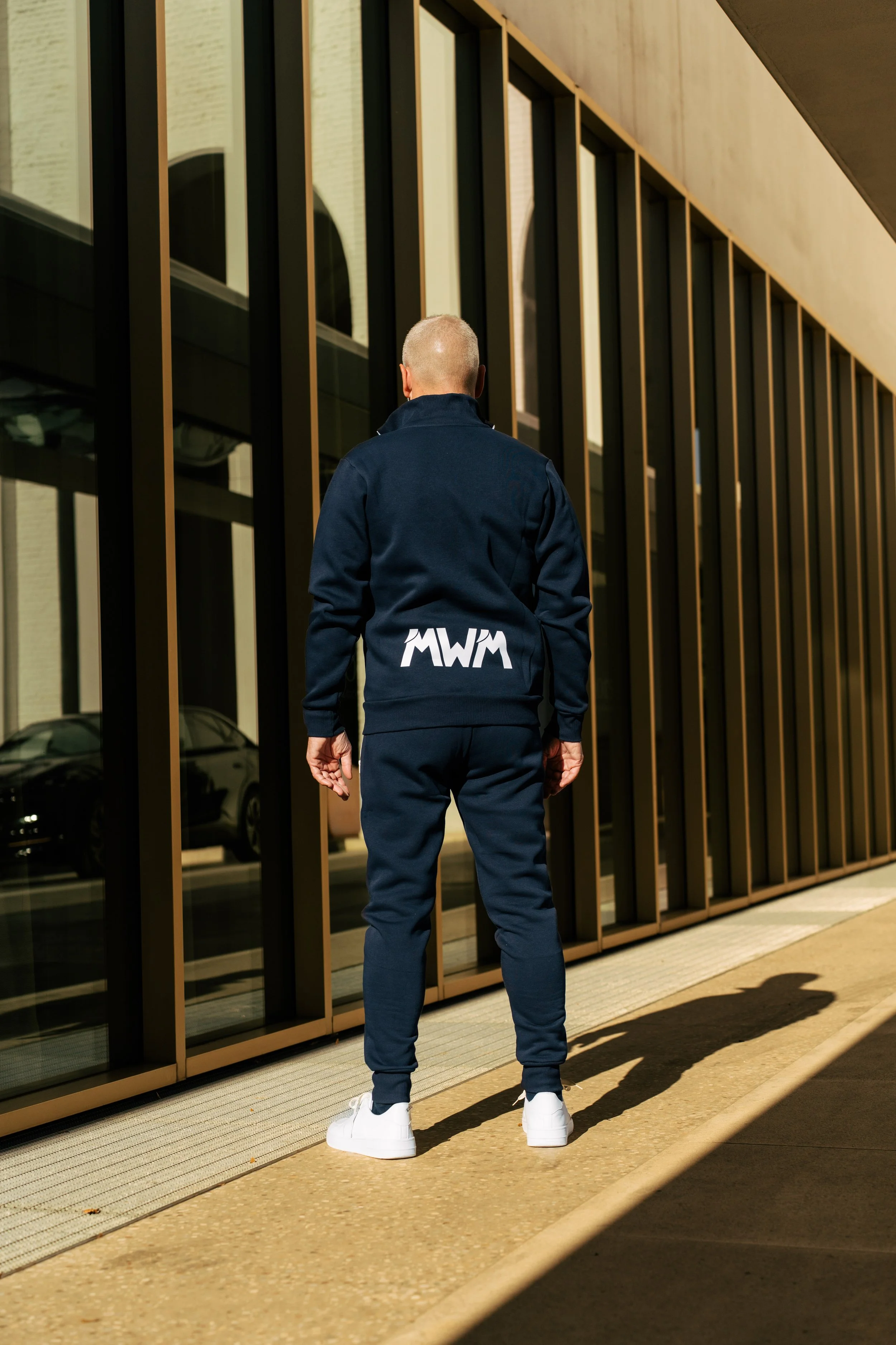 Uomo vestito con abbigliamento sportivo blu, con scritta 'MWM' sulla schiena, che cammina accanto a un edificio con grandi vetrate.
