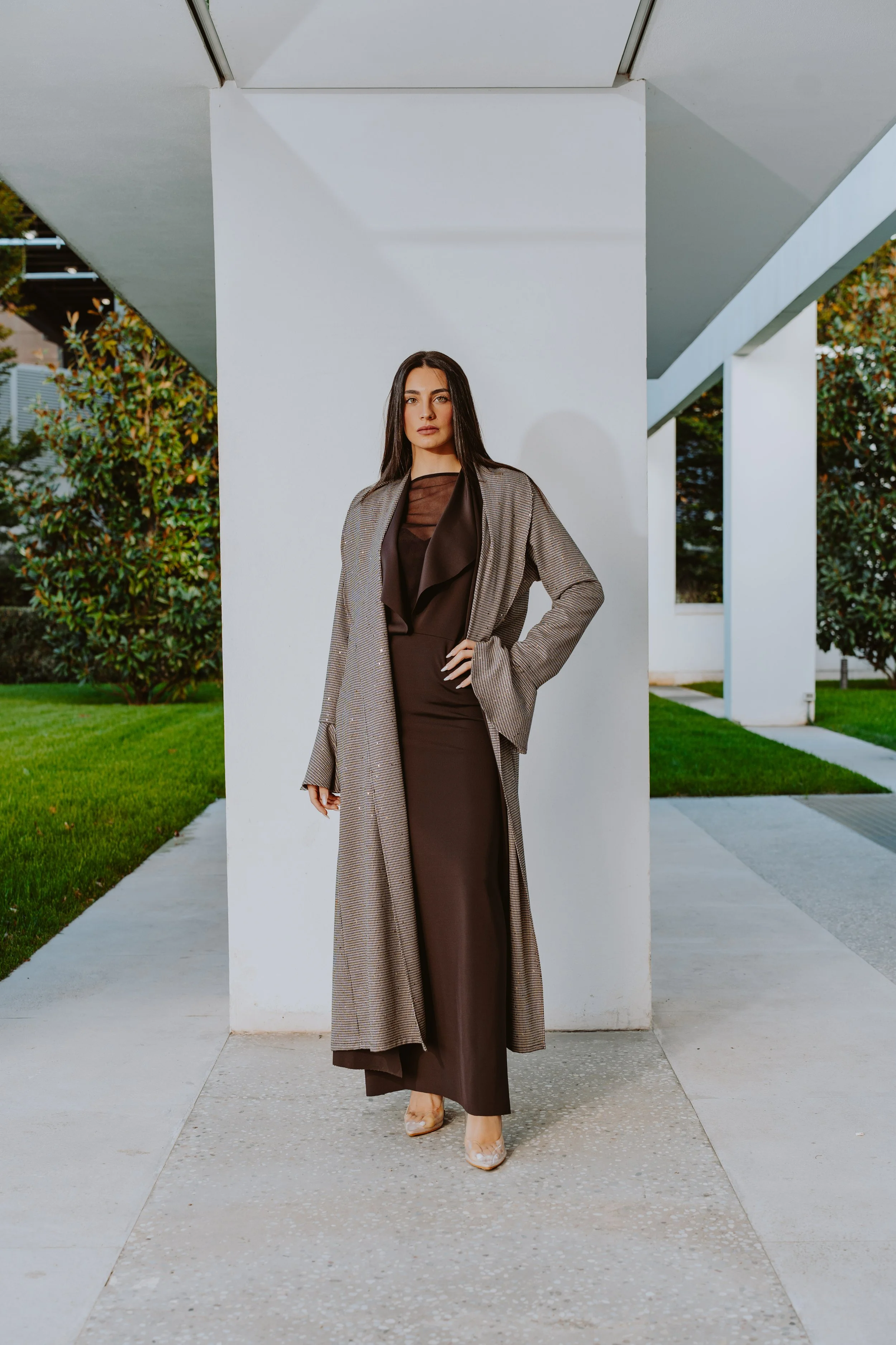 Una donna indossa un abito lungo marrone e un cappotto a quadri, si trova davanti a un muro bianco all'aperto con un contorno di vegetazione verde.