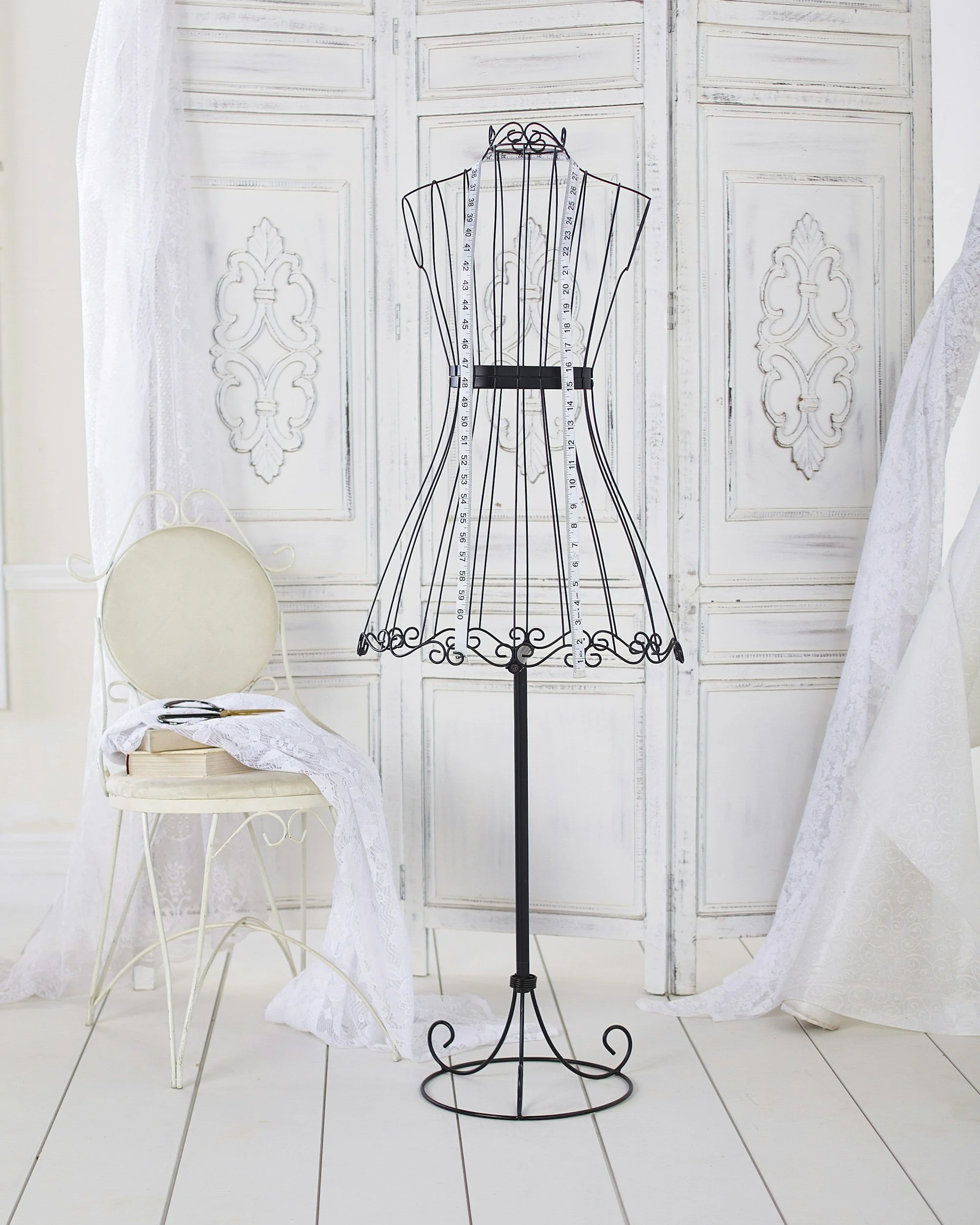 Un manichino in ferro con misuratori appesi, che si trova tra una sedia bianca con un drappo di pizzo bianco sopra e delle tende di pizzo bianco alle estremità. Lo sfondo è di pannelli di legno dipinti di bianco con dettagli decorativi.