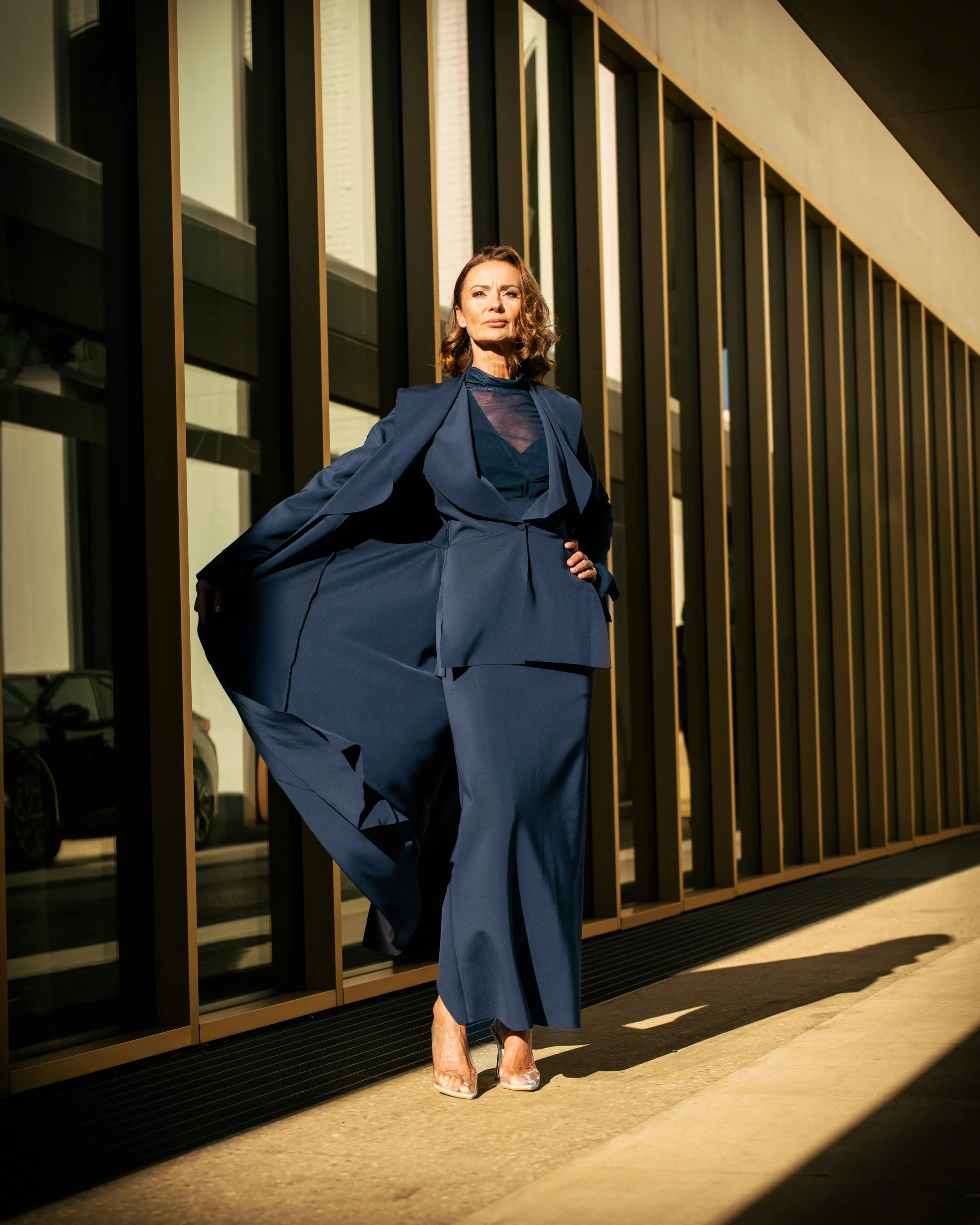 Donna in completo blu con giacca lunga e pantaloni ampi, posa davanti a un edificio moderno con parete di vetro e pannelli dorati, con luce solare che crea ombre sul pavimento.