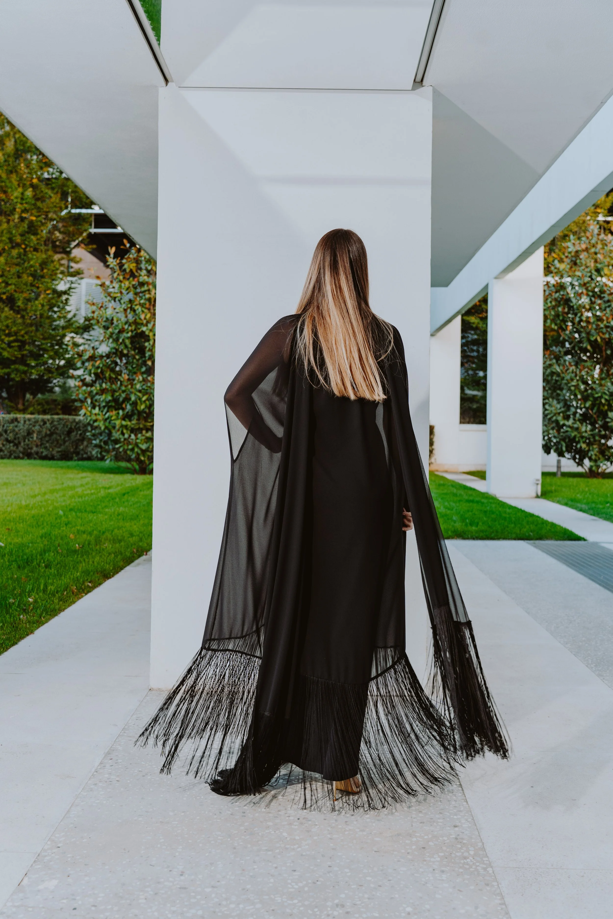 Una donna con lunghi capelli castani vestita con un abito nero, vista da dietro, che si trova in un'area moderna con pareti bianche e grandi finestre, circondata da vegetazione verde.