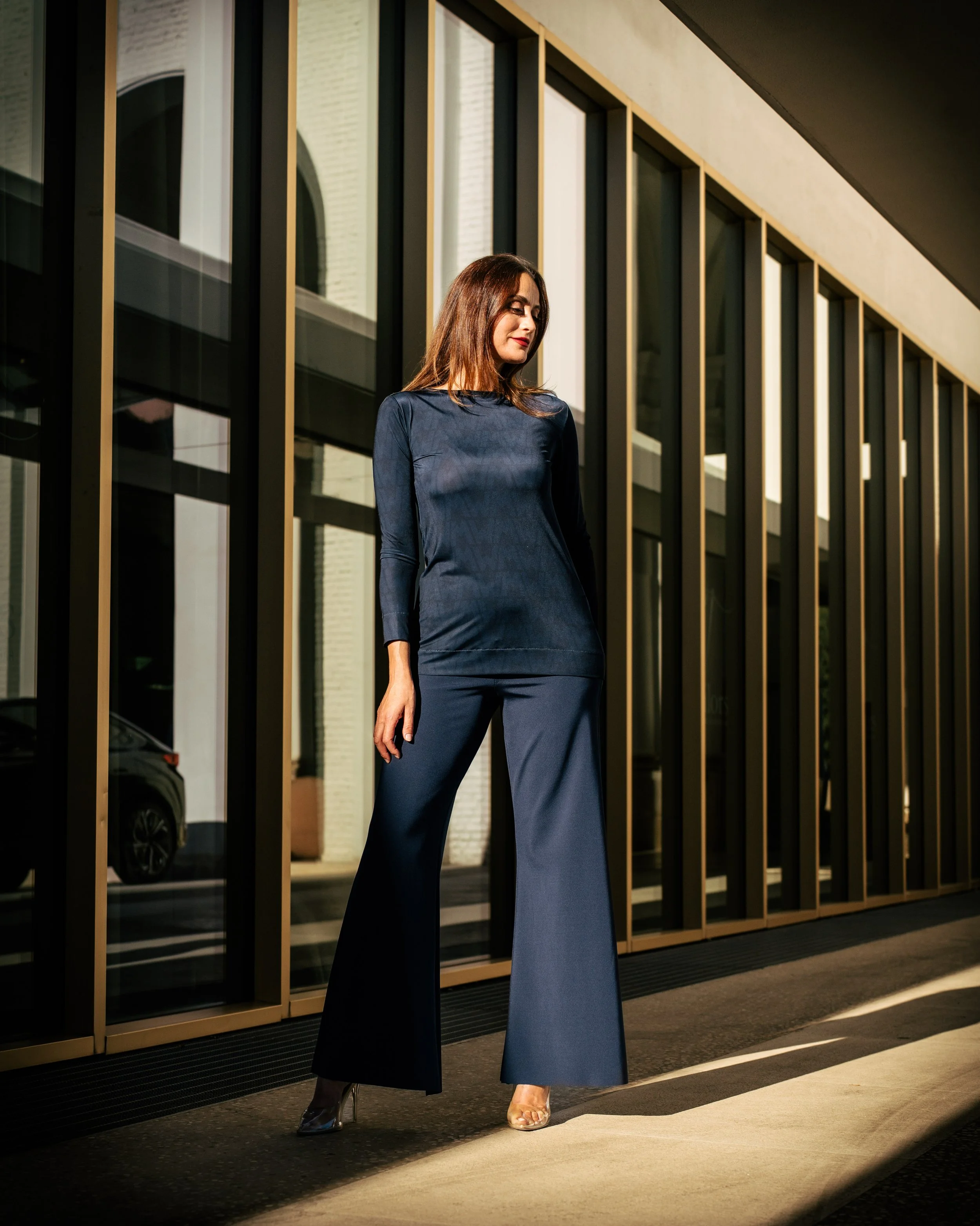 Donna elegante in abito blu scuro e pantofole alte, cammina davanti a un edificio con grandi finestre, illuminata dalla luce solare.