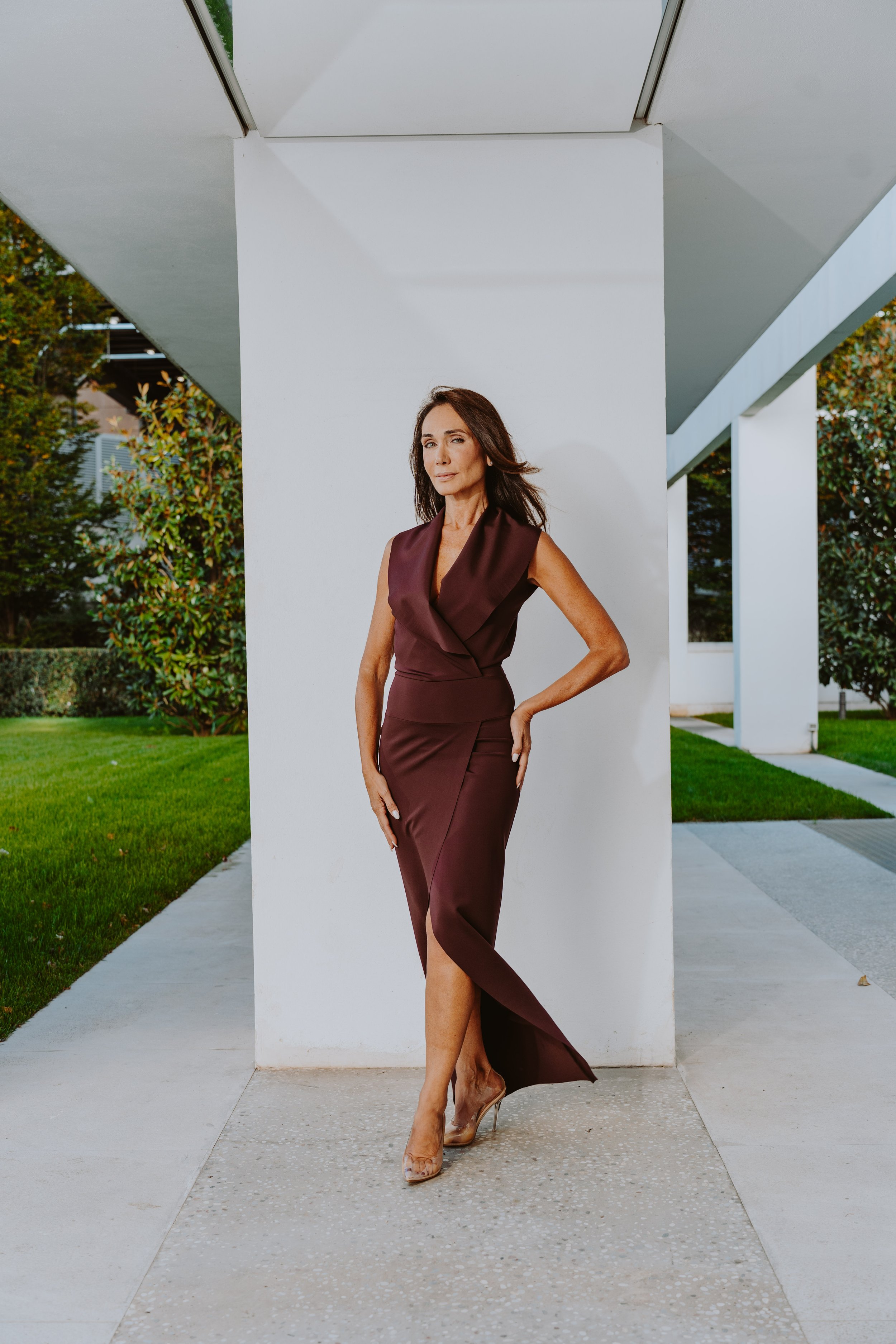 Una donna elegante indossa un vestito lungo color borgogna con tacchi trasparenti, si trova davanti a un muro bianco in un ambiente esterno moderno.
