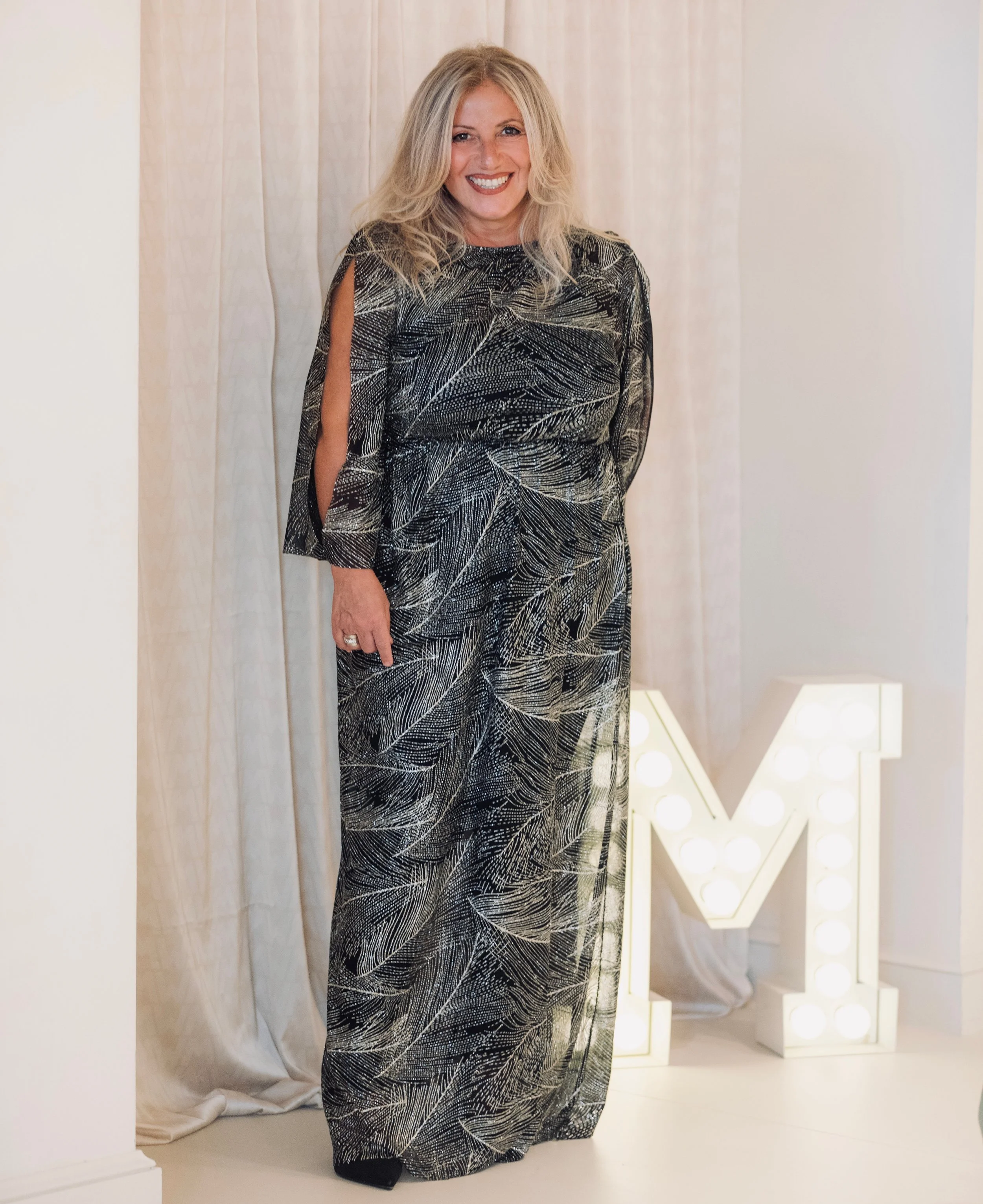 Donna sorridente in abito lungo con stampa a foglie, accanto a una lettera luminosa M.