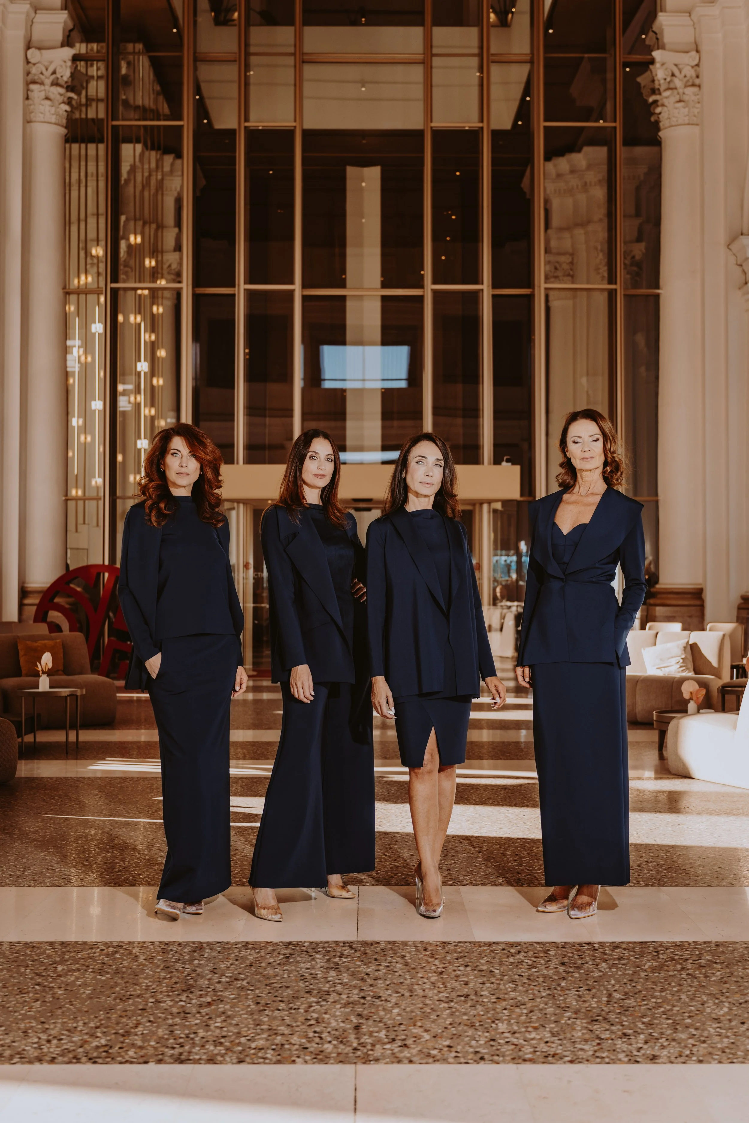 Quattro donne vestite elegantemente in abiti blu scuro posano in un grande atrio con archi e colonne.