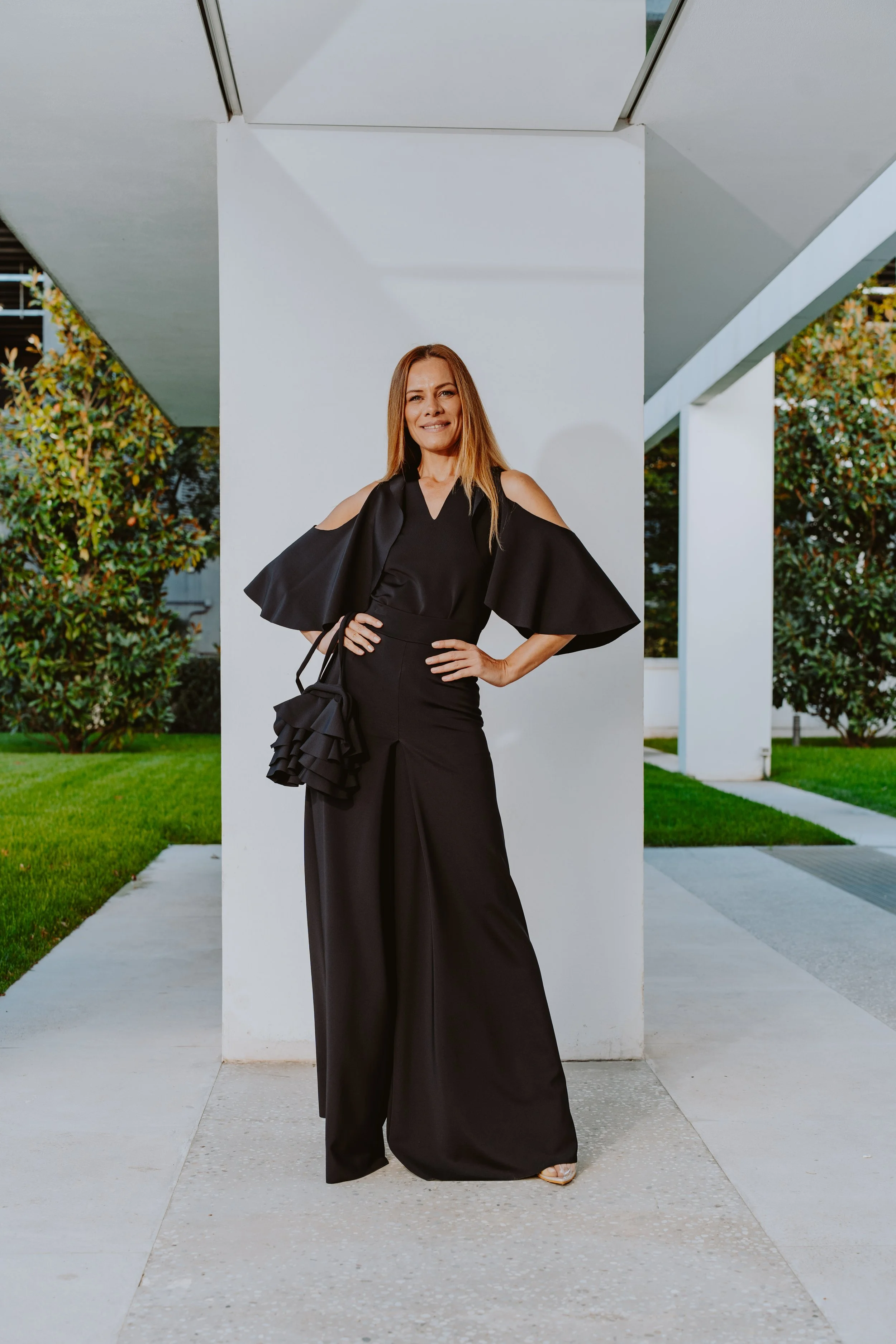 Una donna in abito nero lungo posa davanti a un muro bianco, con un cortile verde e un edificio moderno sullo sfondo.