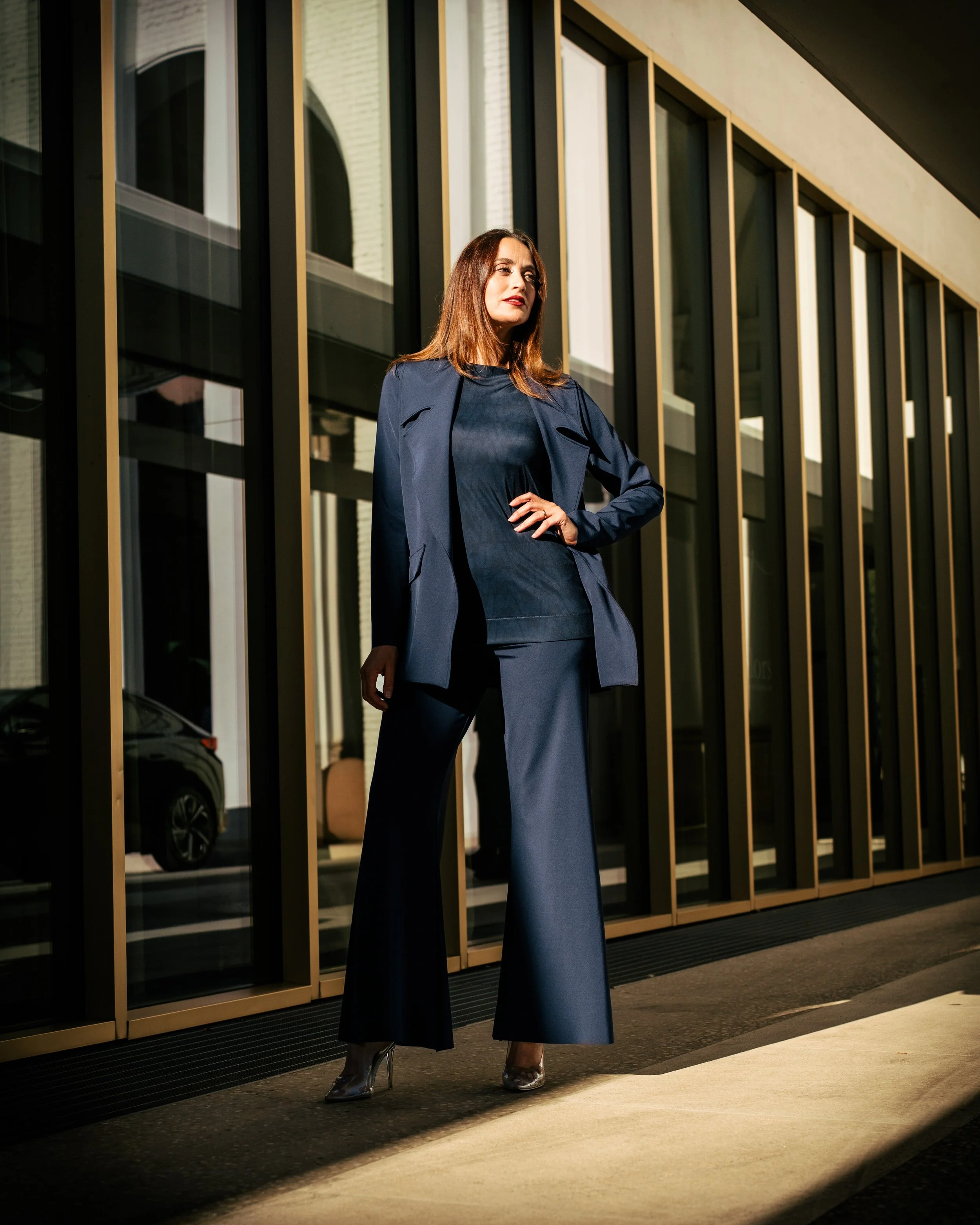 Donna che indossa un completo blu scuro con pantaloni larghi che si trova davanti a un edificio moderno con pannelli di vetro e metallo.