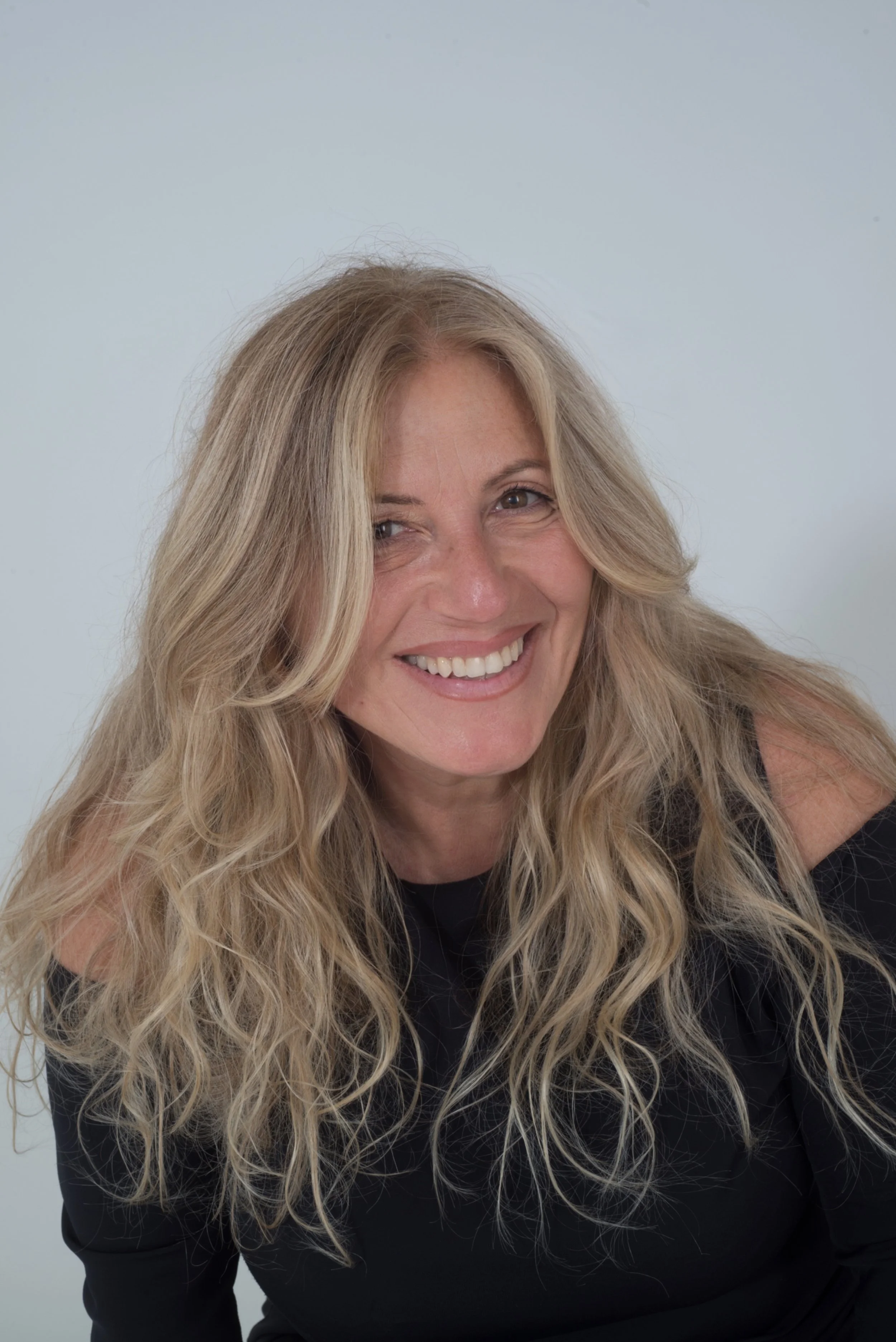 Una donna sorridente con capelli biondi lunghi e ondulati, indossa un top nero con spalle scoperte, sfondo bianco