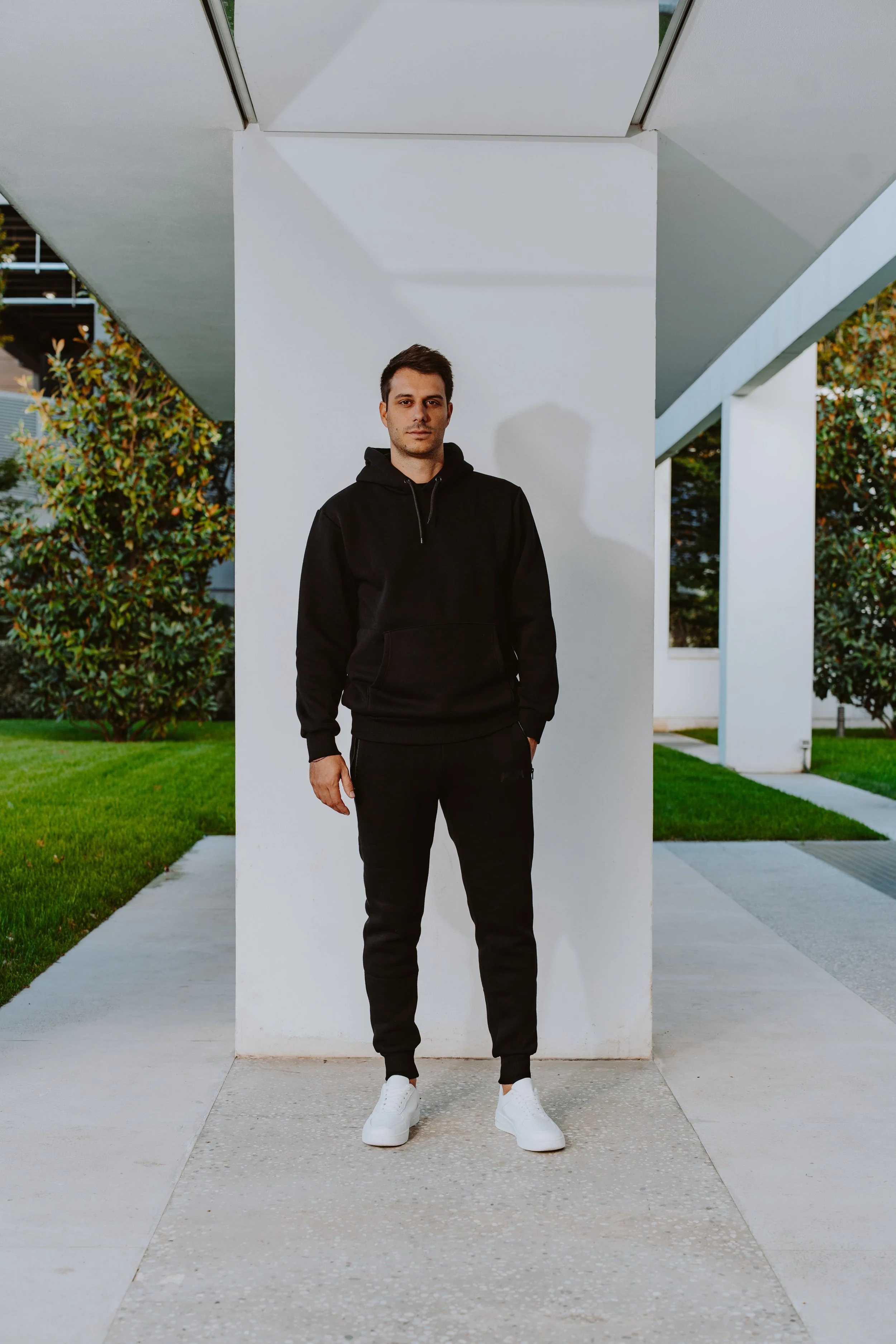 Un uomo vestito casualmente in tuta nera con scarpe bianche si trova davanti a una colonna bianca in un ambiente urbano con un edificio moderno e vegetazione sullo sfondo.