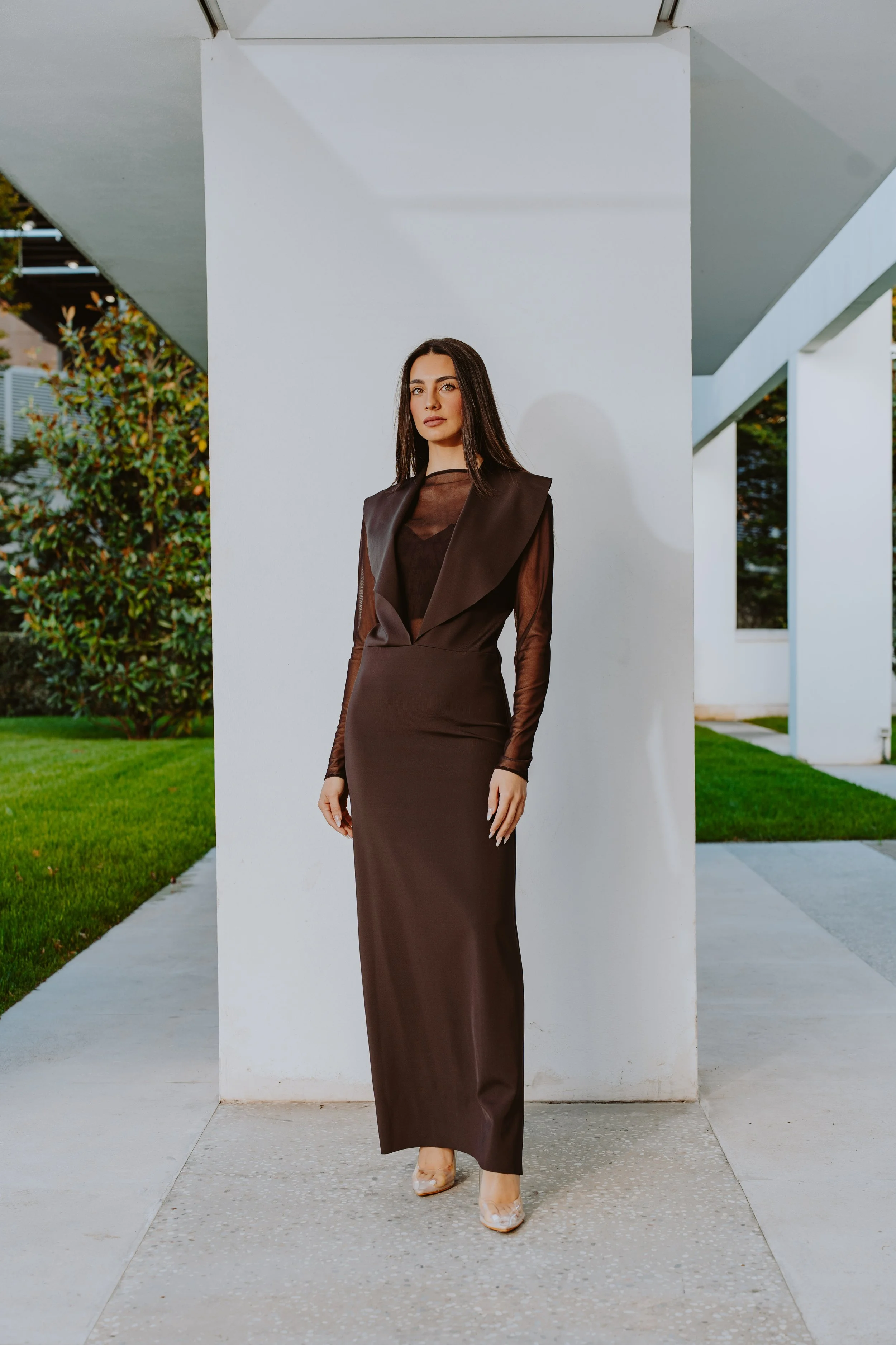 Donna in abito lungo marrone con trasparenze, posa davanti a un muro bianco in un'esterno moderno.