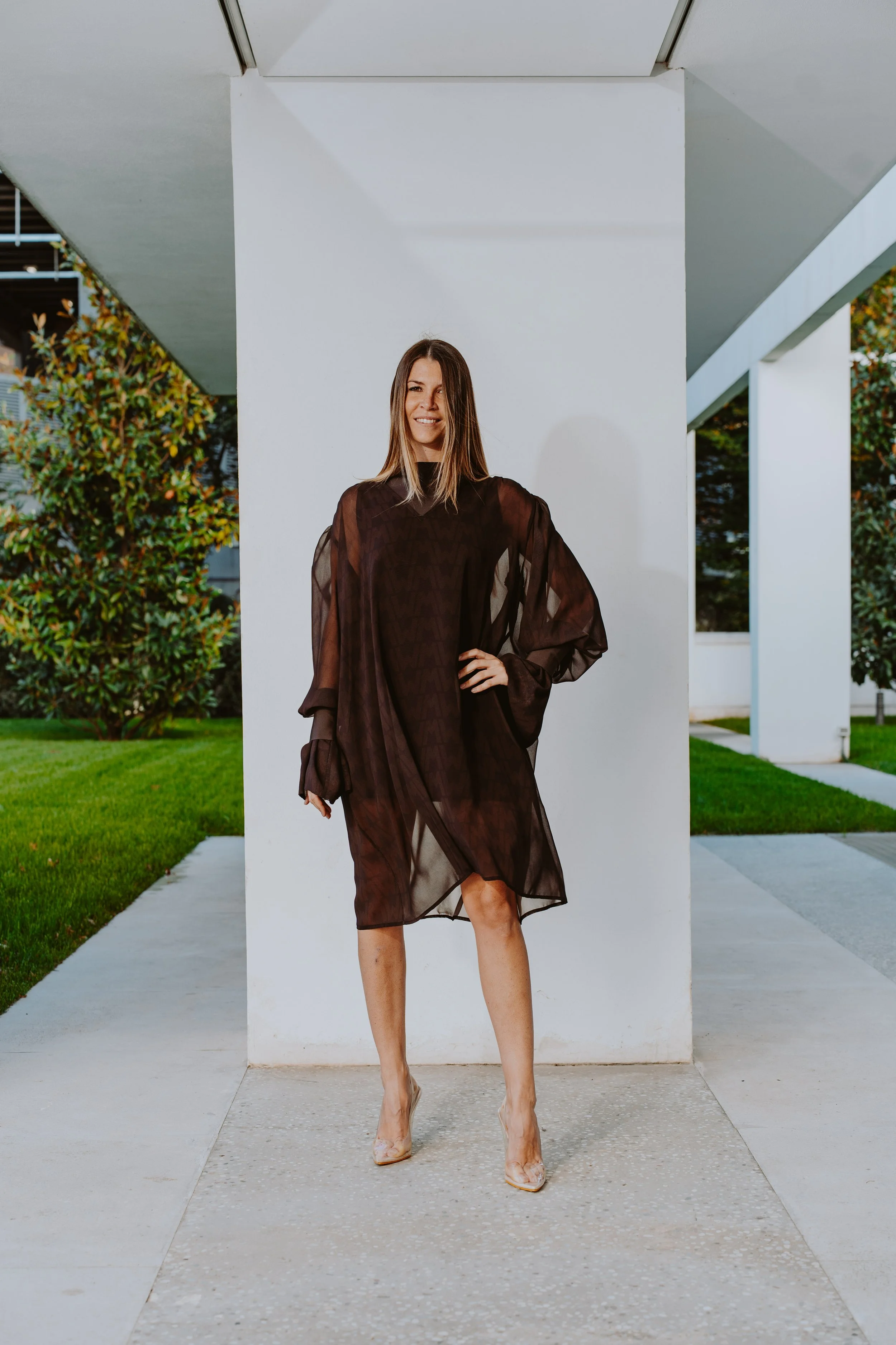 Una donna vestita con un abito nero trasparente davanti a una parete bianca, con un paesaggio di edifici e alberi sullo sfondo.