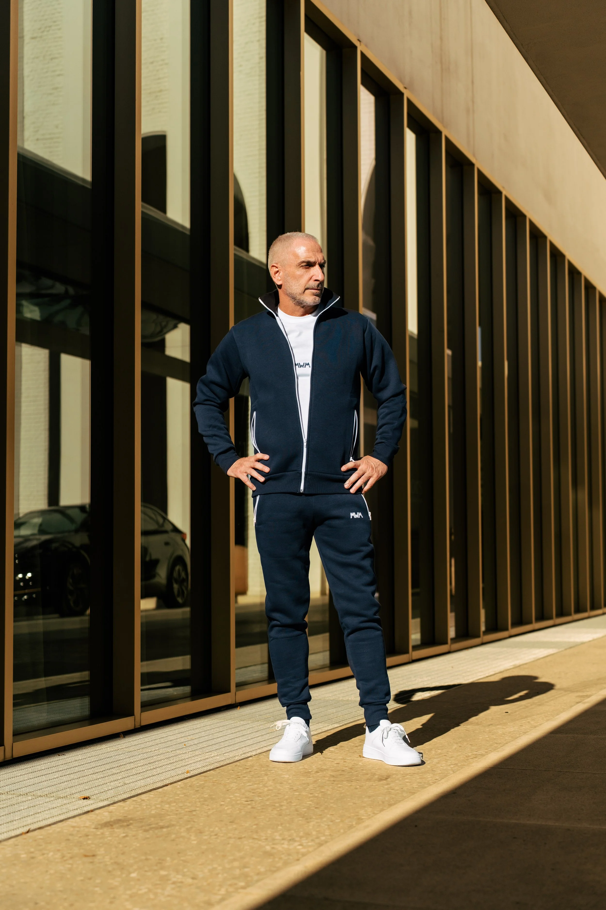 Uomo che indossa abbigliamento sportivo bianco e blu, camminando davanti a un edificio moderno con grandi vetrate.