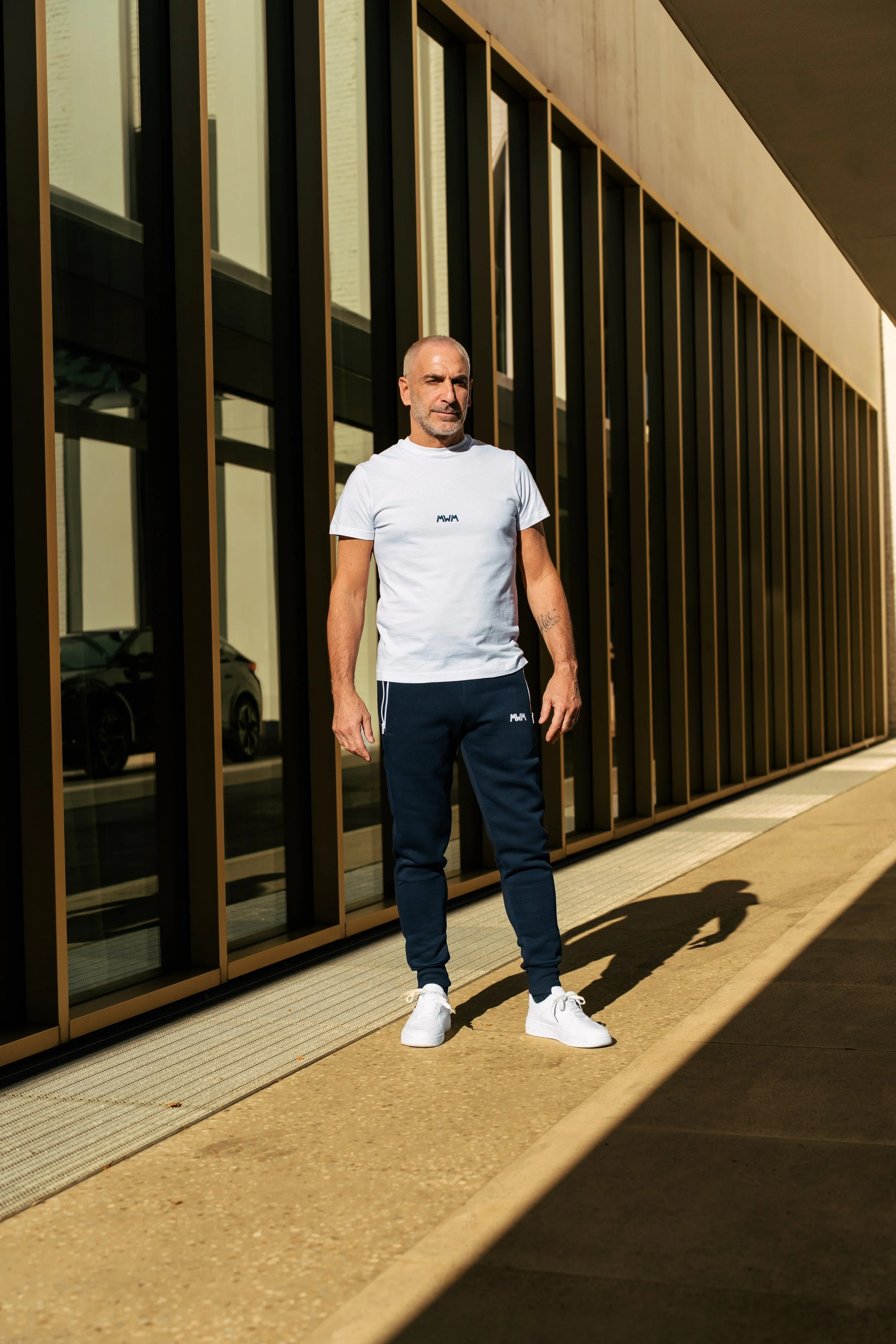 Uomo adulto in abbigliamento sportivo con pantaloni jogger e maglietta bianca, che si trova all'aperto davanti a un edificio con pareti di vetro e struttura metallica, con ombra proiettata sul pavimento.