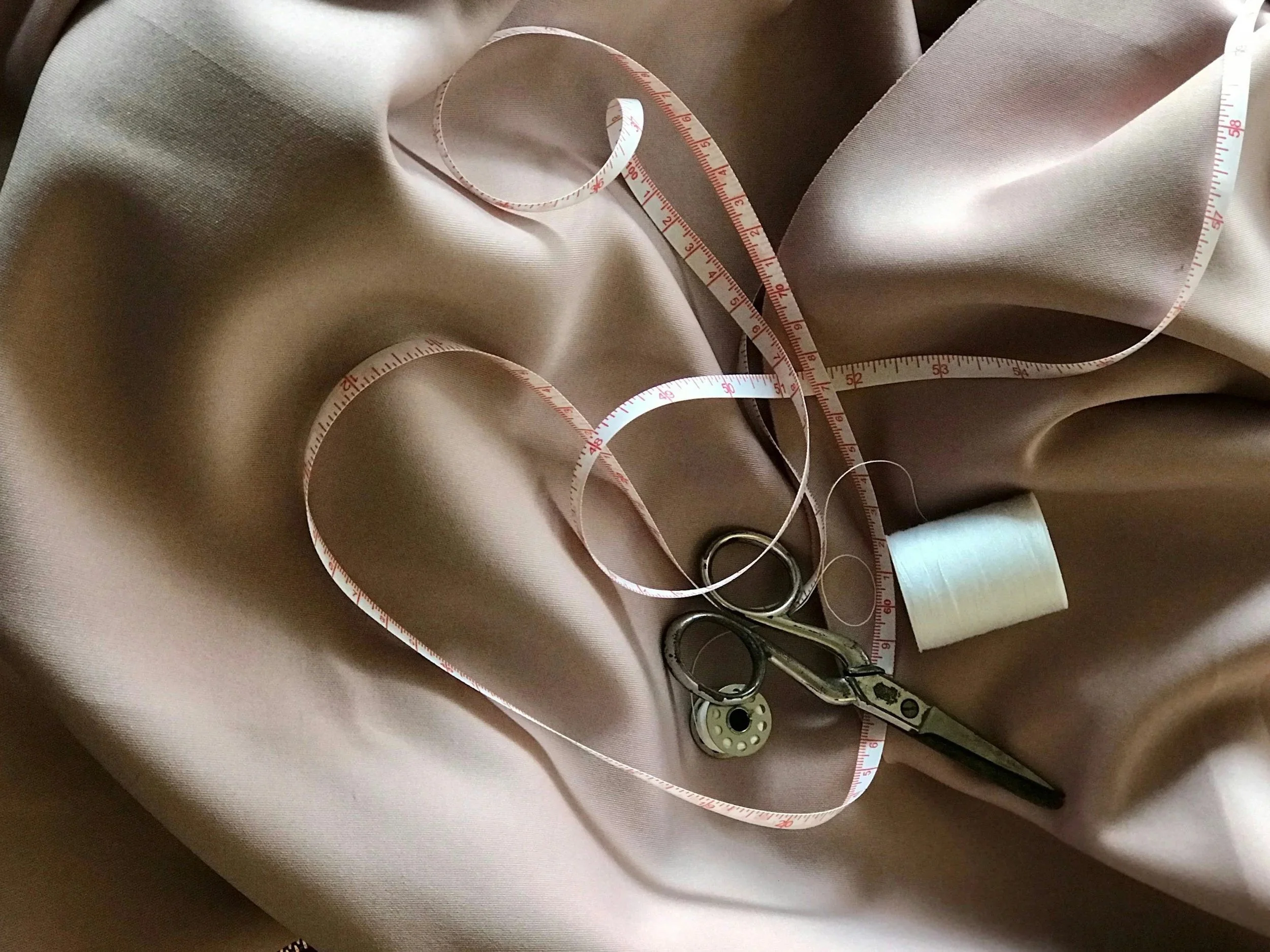 Tessuto beige, forbici, metro a nastro, filo e spoletta per cucire su tessuto.