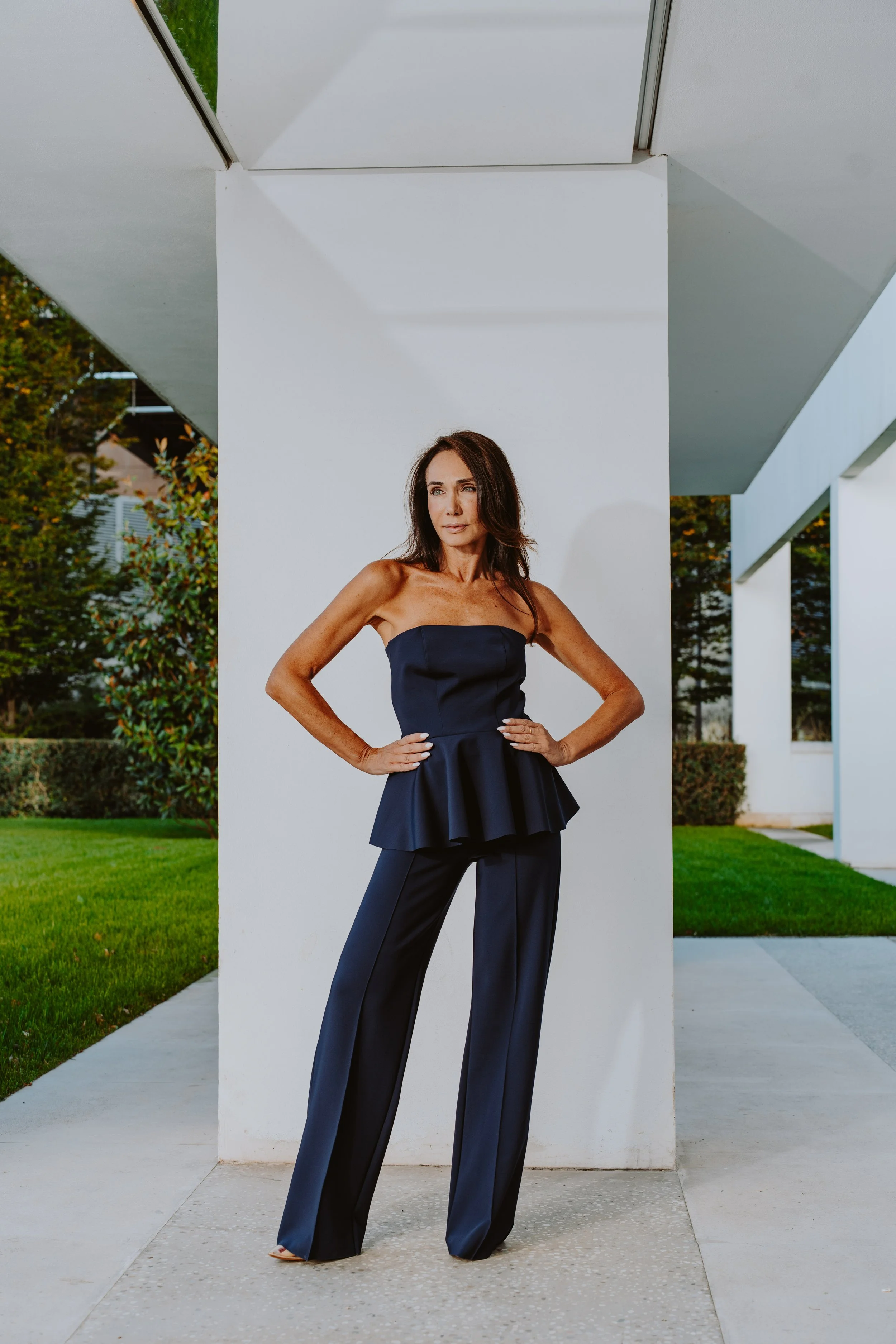 Donna con abito nero elegante, posando all'aperto davanti a una struttura moderna bianca, con vegetazione in background.