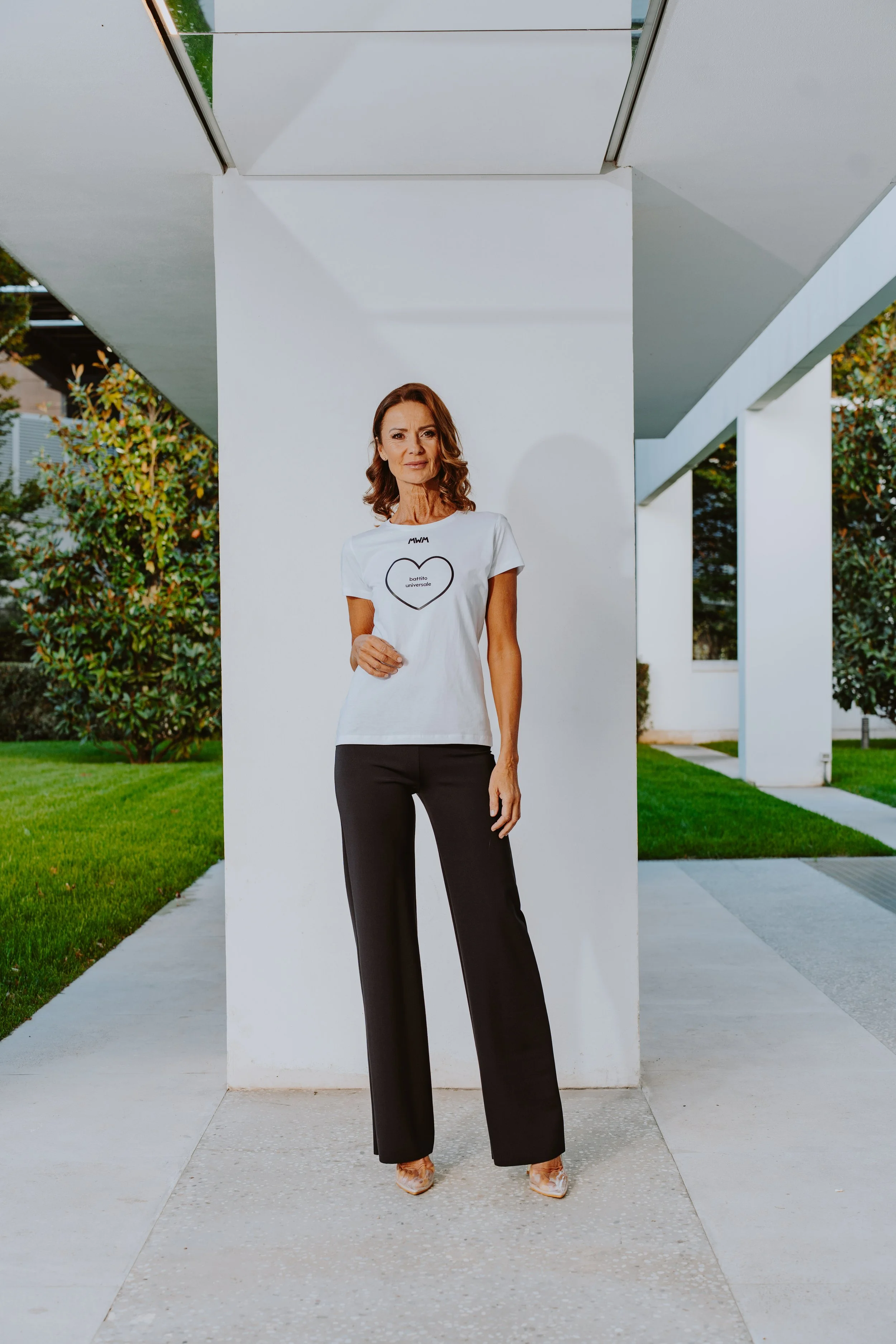Donna con camicia bianca e pantaloni neri che si trova davanti a una parete bianca, con un'area di verde e edifici moderni sullo sfondo.