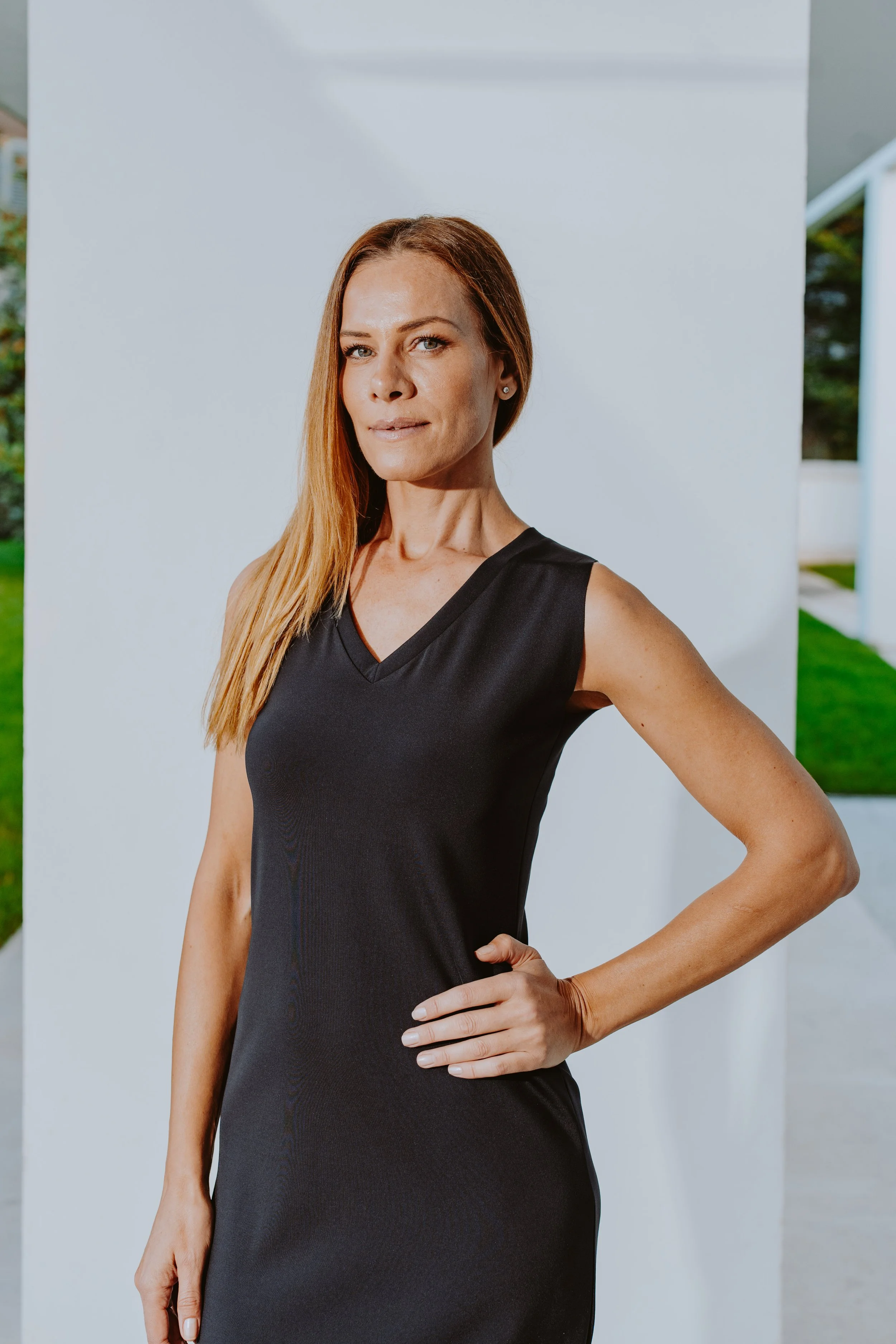 Donna con vestito nero, posa con mano sulla naturale curva del fianco, faccia seria, sfondo moderno con parete bianca e spazi verdi.