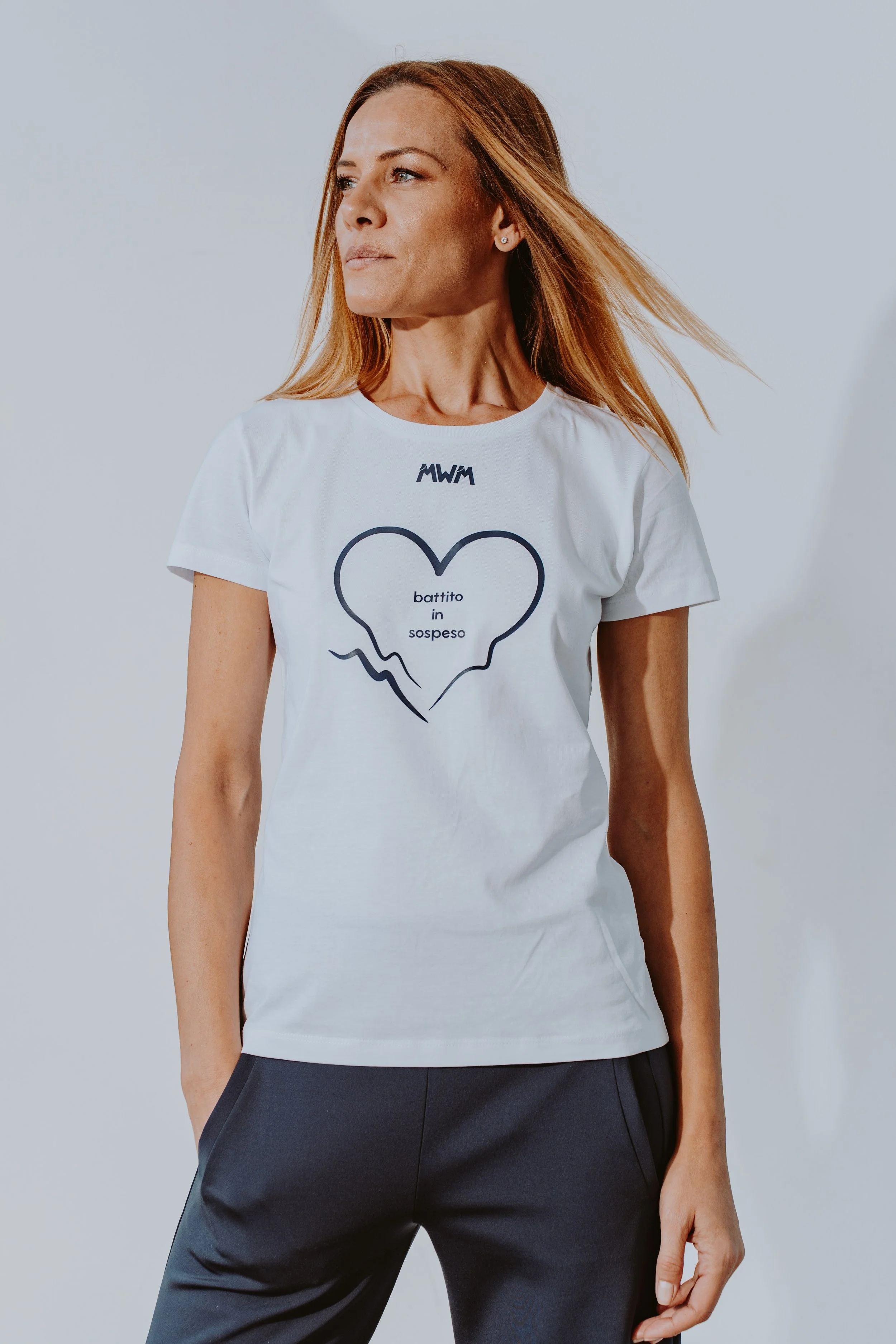Donna con capelli castani e corti indossa una maglietta bianca con una scritta e un'immagine di un cuore spezzato, e pantaloni scuri.