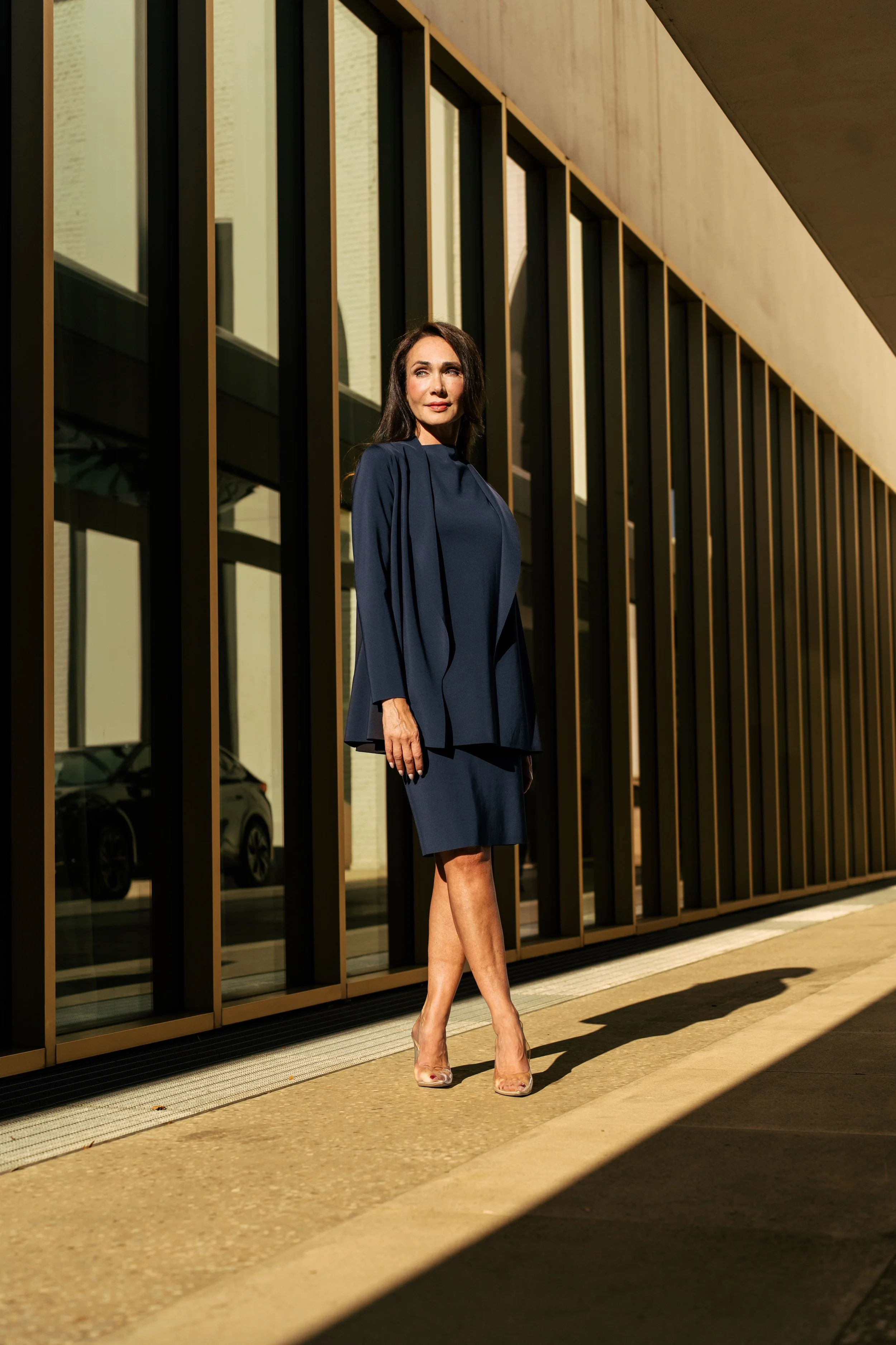 Una donna vestita con un abito blu scuro si trova davanti a un edificio con grandi finestre e montanti dorati, illuminata dalla luce del sole.