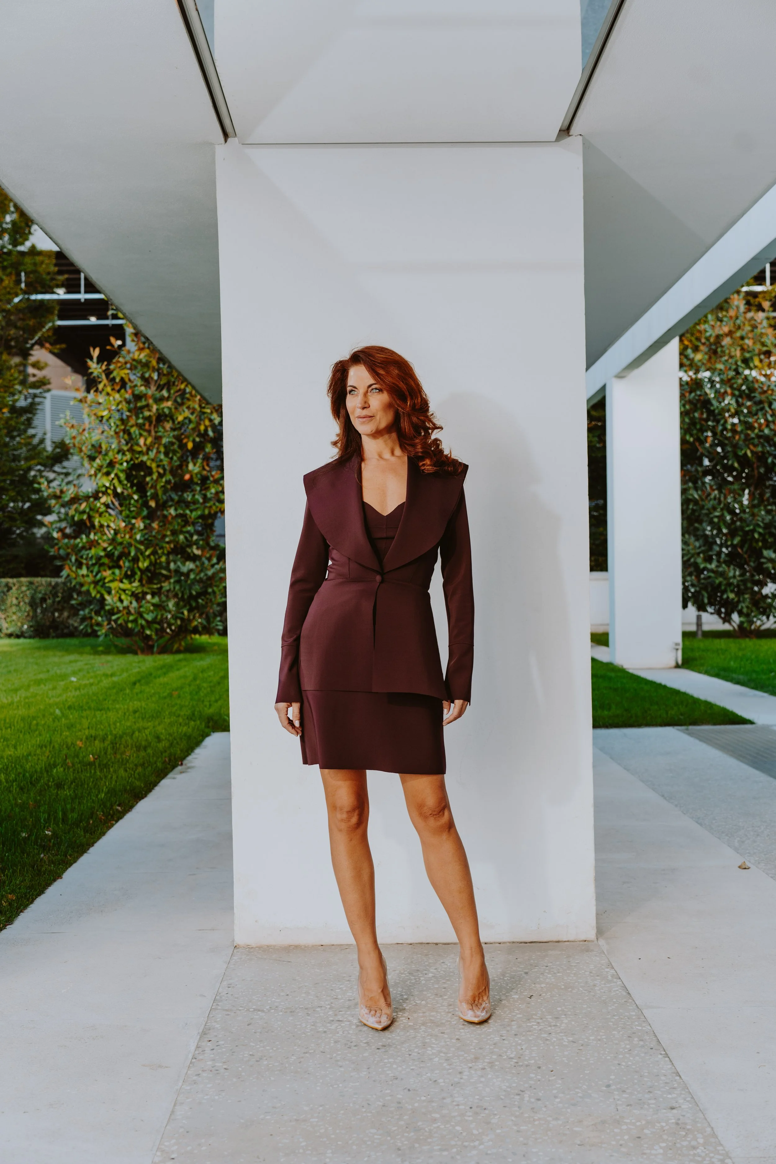 Donna con abito da sera vestita di bordeaux, che si staglia contro un muro bianco, con angoli di edifici moderni e vegetazione verde sullo sfondo.