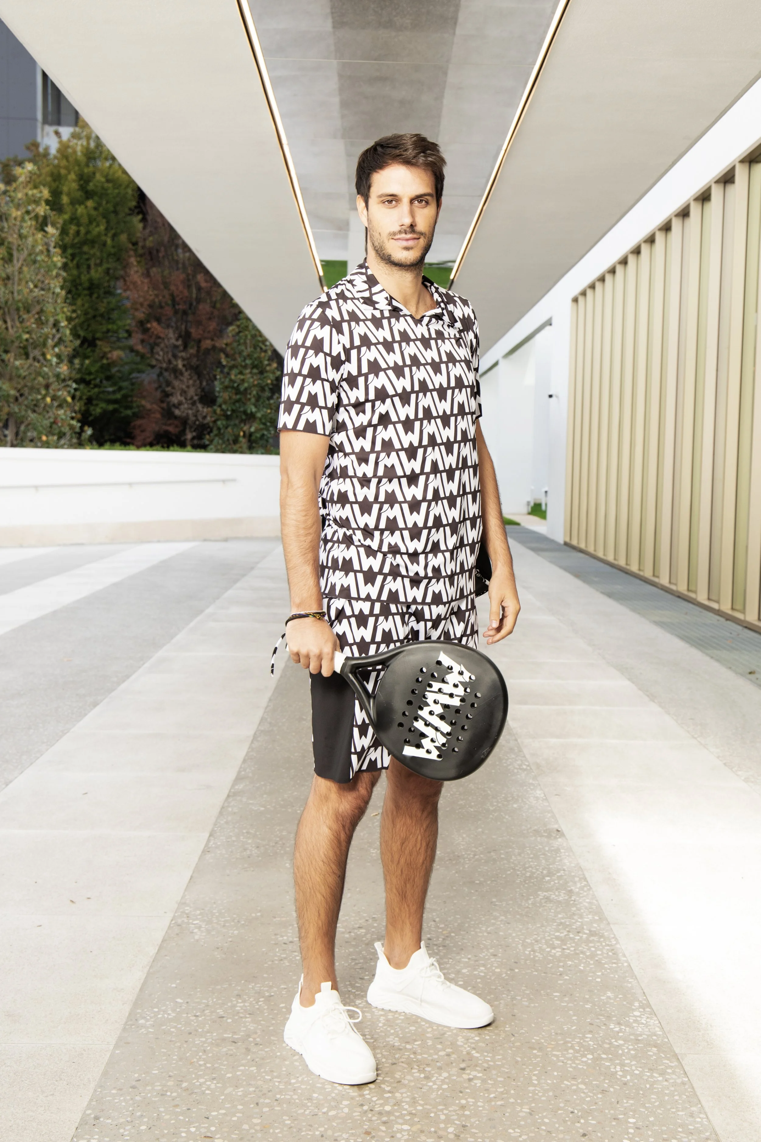 Un uomo giovane con vestiti fantasia bianco e nero tiene una racchetta da paddle in mano, in piedi all'aperto in un'area moderna.