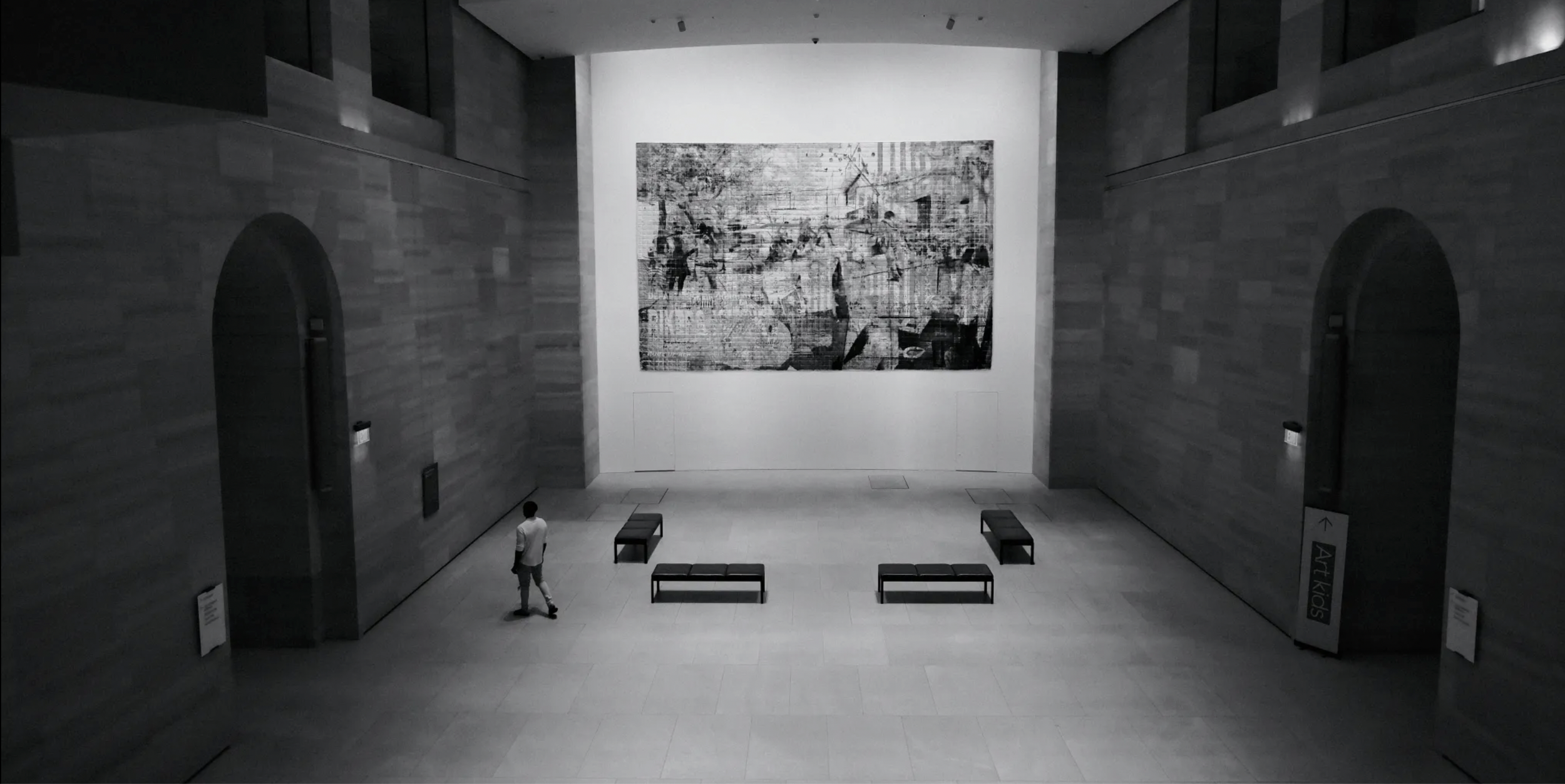 A person walking in an art museum with gray walls and benches, looking at a large black-and-white abstract painting on the wall.