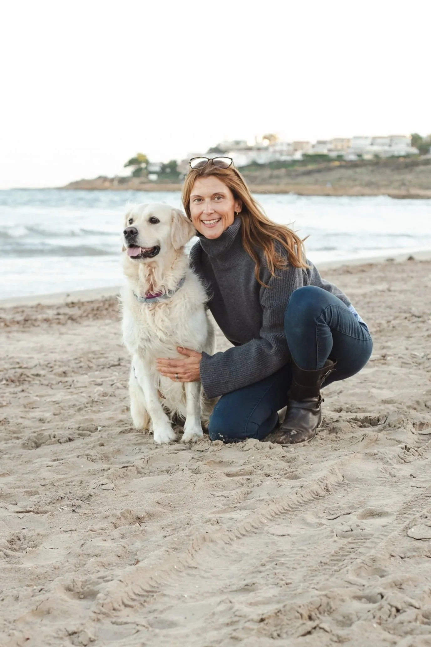 A relaxed, hwoman  on a sandy beach, smiling, with her arm around a large golden retriever dog. In the background, there are ocean waves and a hillside with houses.