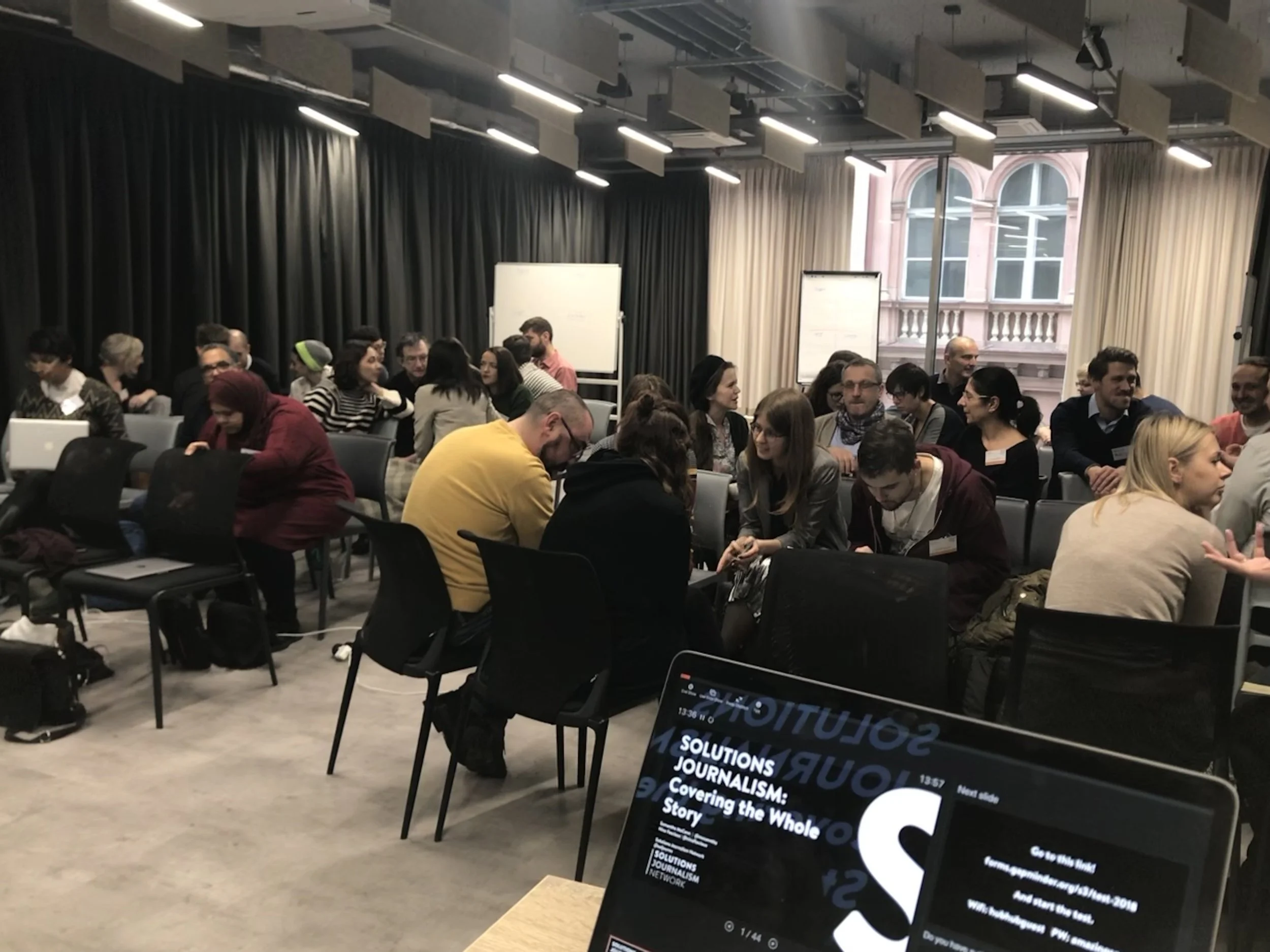 A conference room filled with people sitting in chairs, engaged in discussions, with large windows and black and beige curtains. There are whiteboards at the front and a laptop displaying a presentation titled 'Solutions Journalism: Covering the Whol