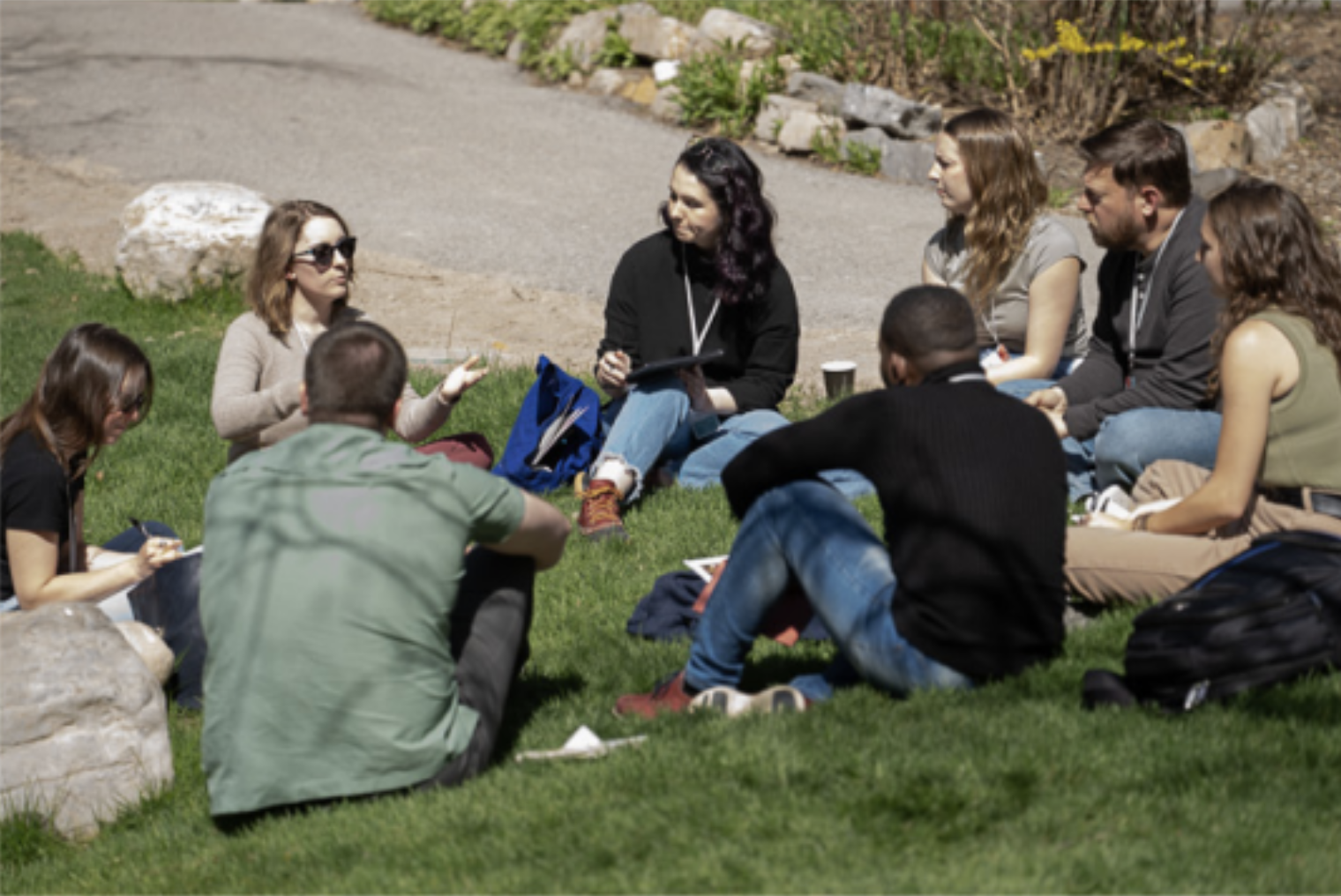 Group of people sitting on grass outside, engaged in a discussion or meeting, some taking notes.