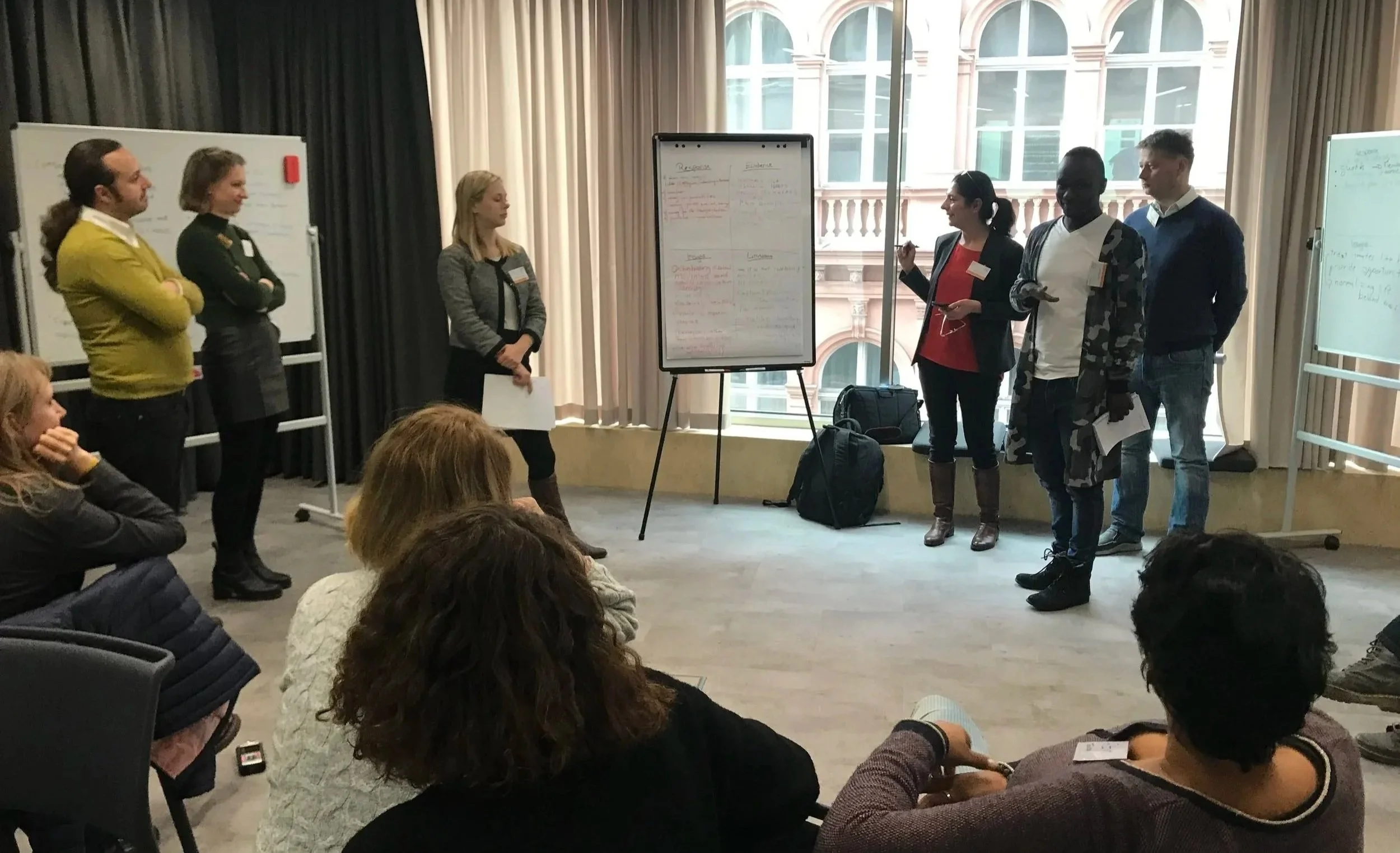 Group of people participating in a workshop or seminar in a conference room, with a presenter pointing at a whiteboard and an audience seated watching.