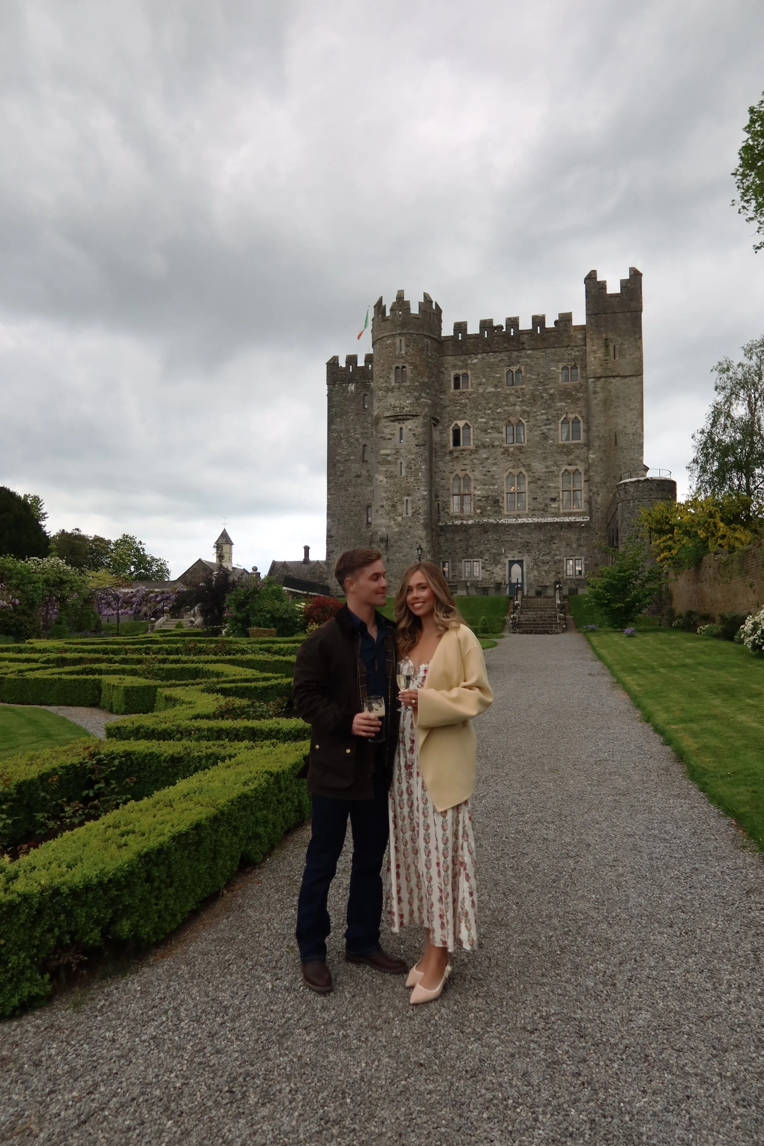A young couple standing on a gravel path in front of a castle, holding glasses of champagne and smiling. The castle has stone walls with towers and battlements, surrounded by well-maintained gardens.