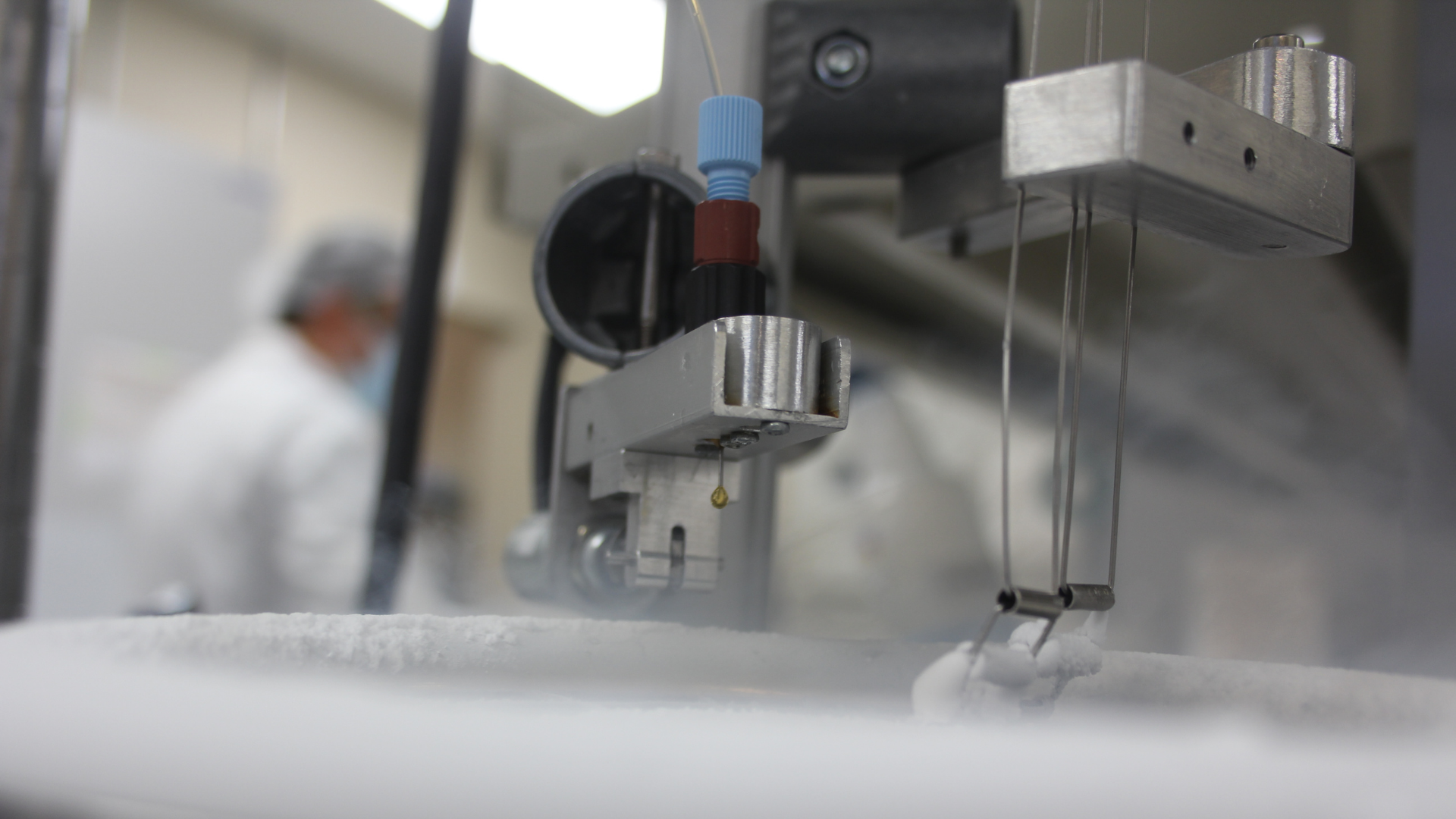 Close-up of medical or laboratory equipment with metal components and wires, with a person wearing protective gear and mask blurred in the background.