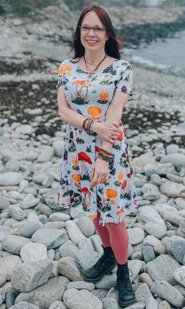 A woman smiling and standing on a rocky beach with a river in the background, wearing a colorful mushroom-themed dress, black boots, and red leggings.