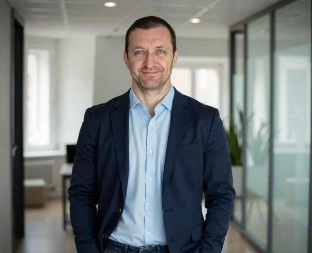 Man in a navy blazer and light blue shirt standing in a modern, glass-enclosed office space.