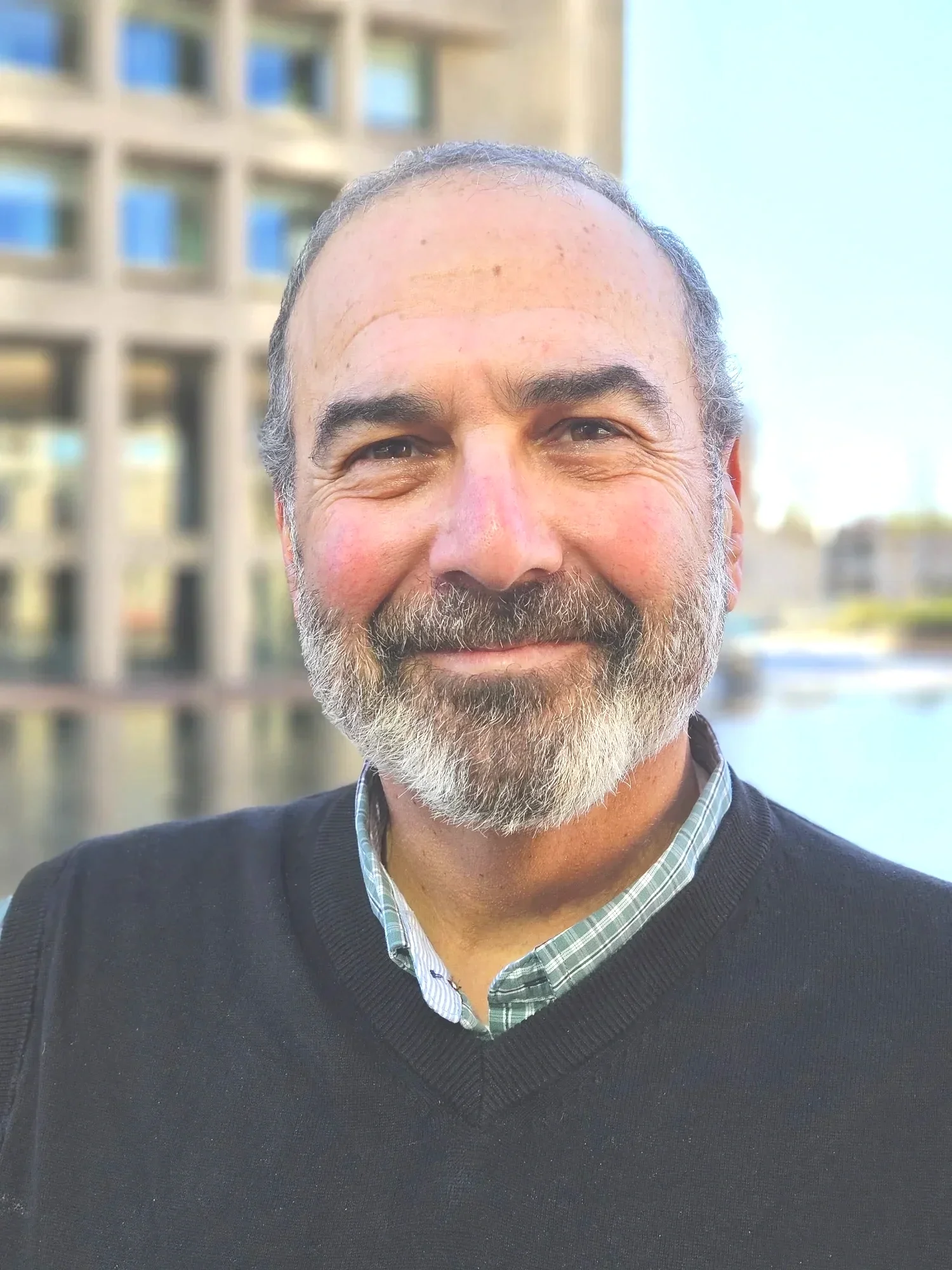 Close-up of a smiling middle-aged man with gray beard and hair, wearing a black sweater over a checkered shirt, outdoors near water with a modern building in the background.
