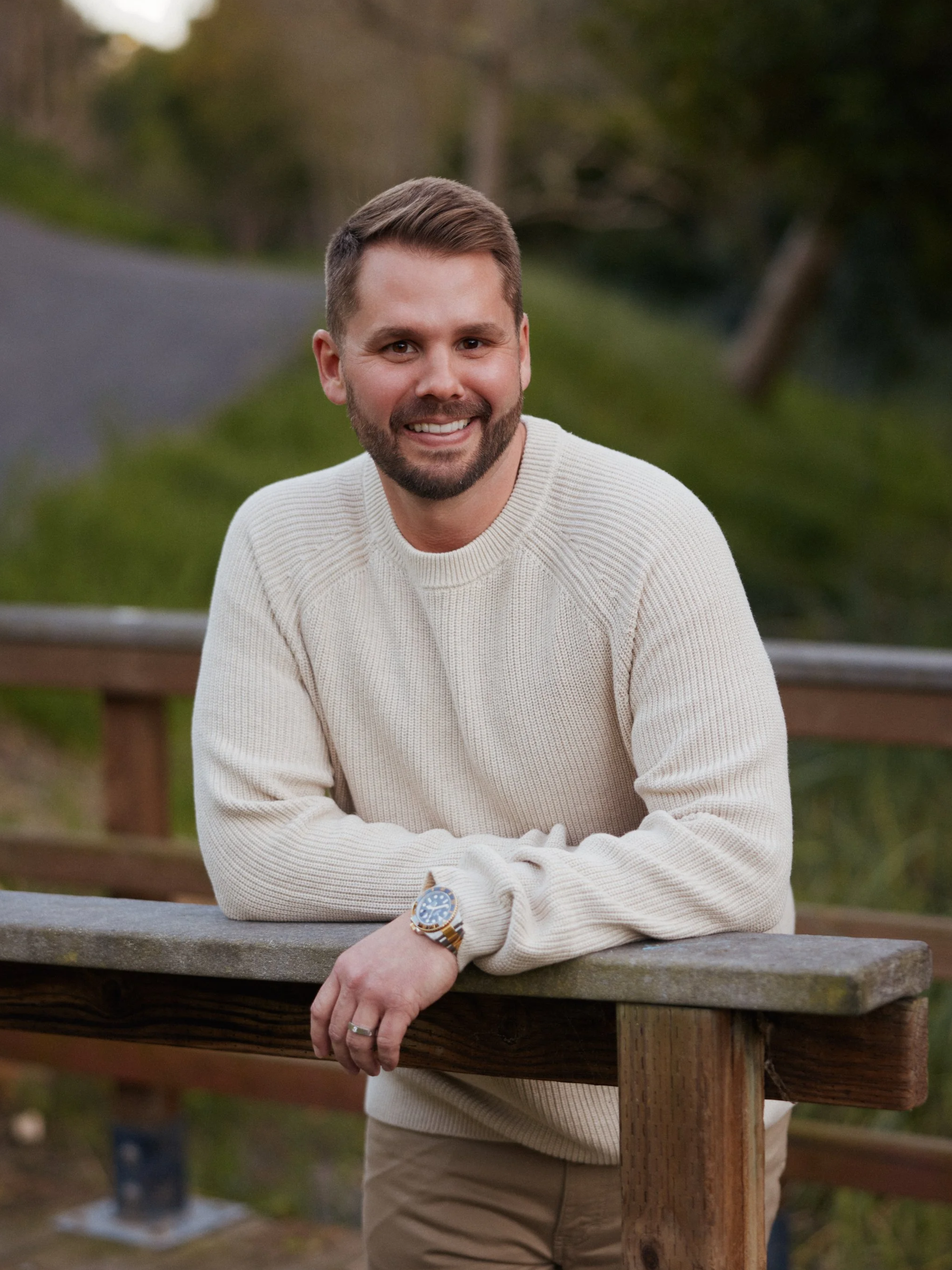 A smiling man with a beard and short hair, wearing a white sweater and a watch, leaning on a wooden railing outdoors.