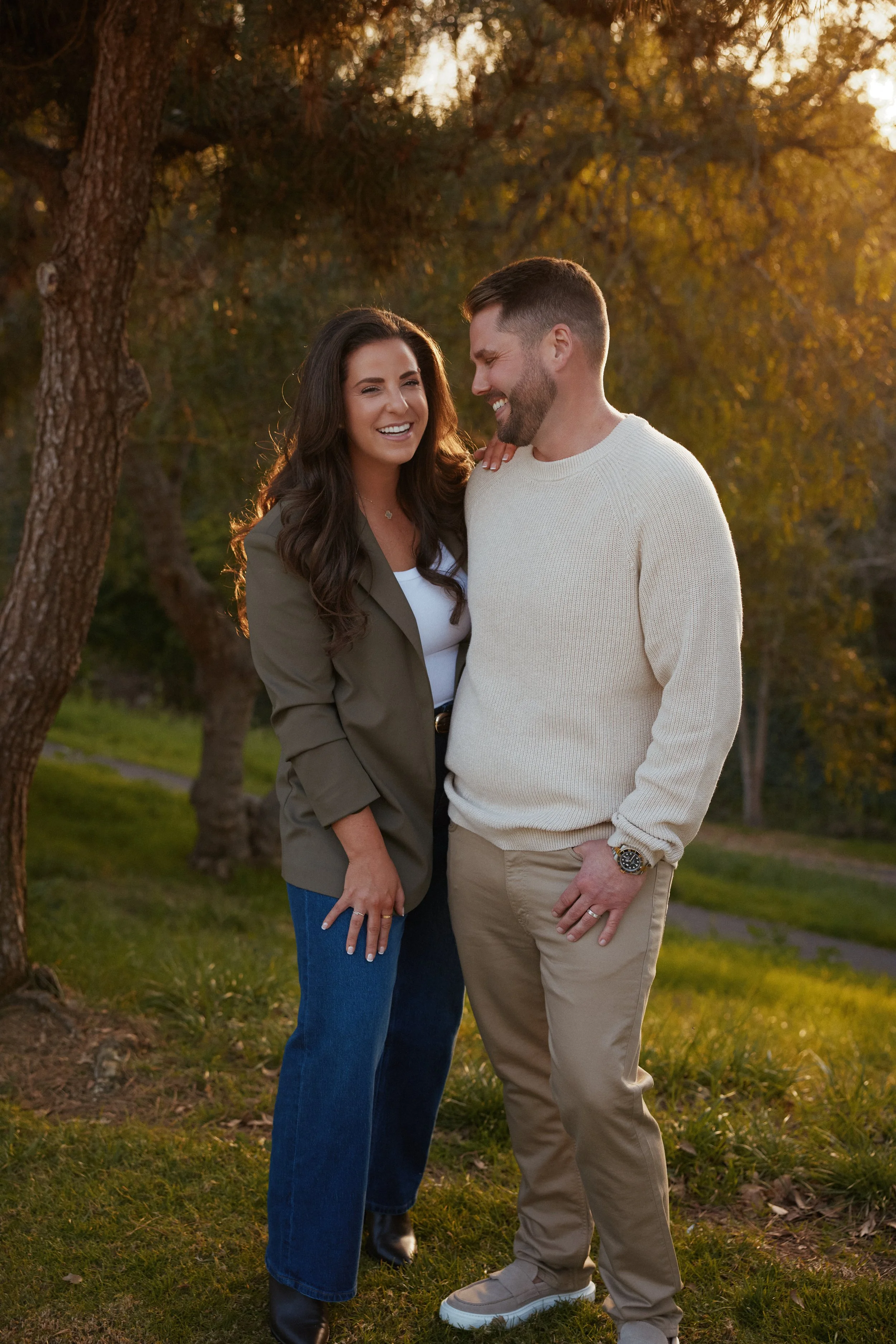 A woman and man are standing outdoors on grass, smiling and laughing together during sunset.