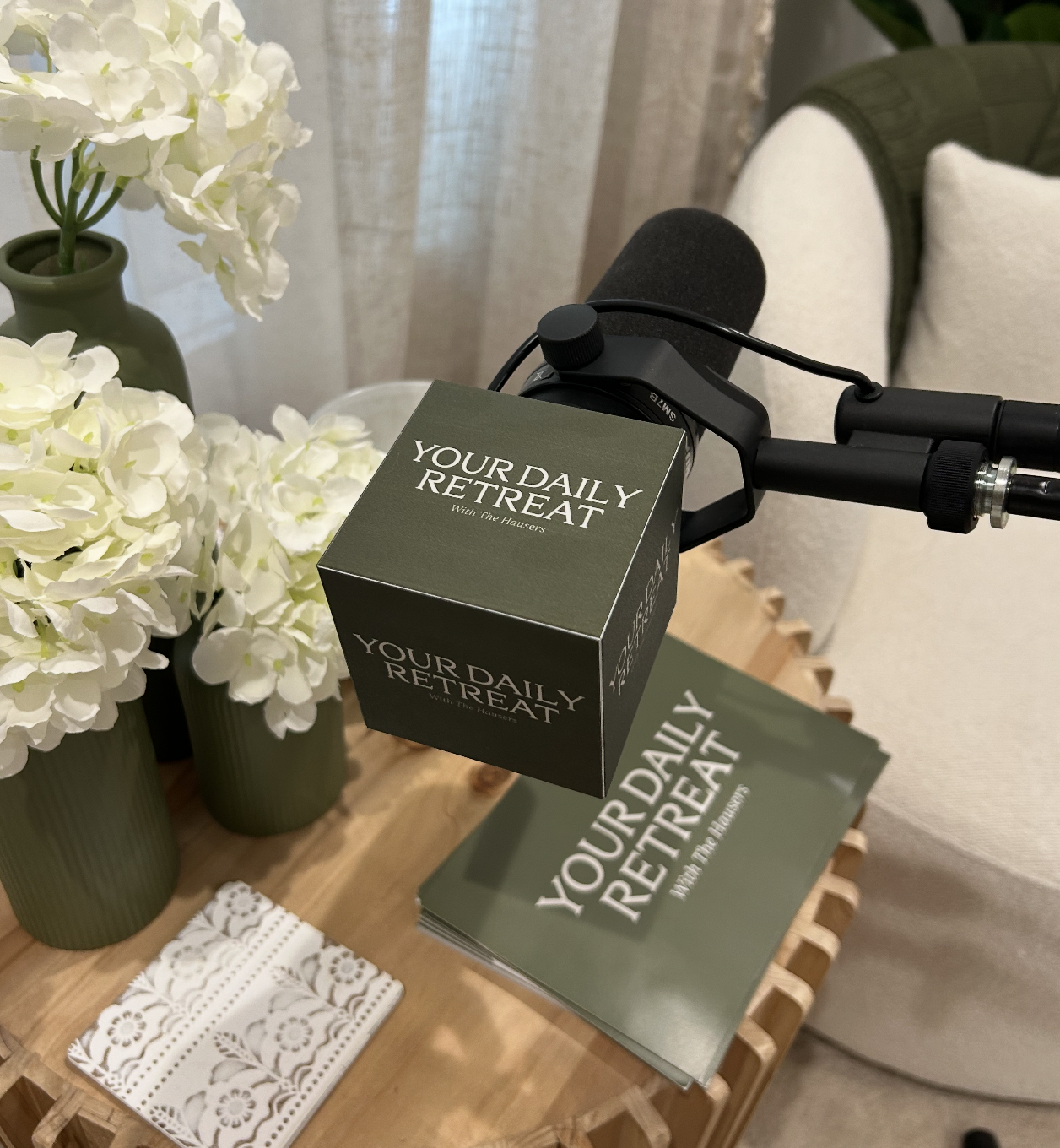 A microphone, a box labeled 'Your Daily Retreat with The Hausers', and a stack of brochures on a wooden table next to vases with white flowers.