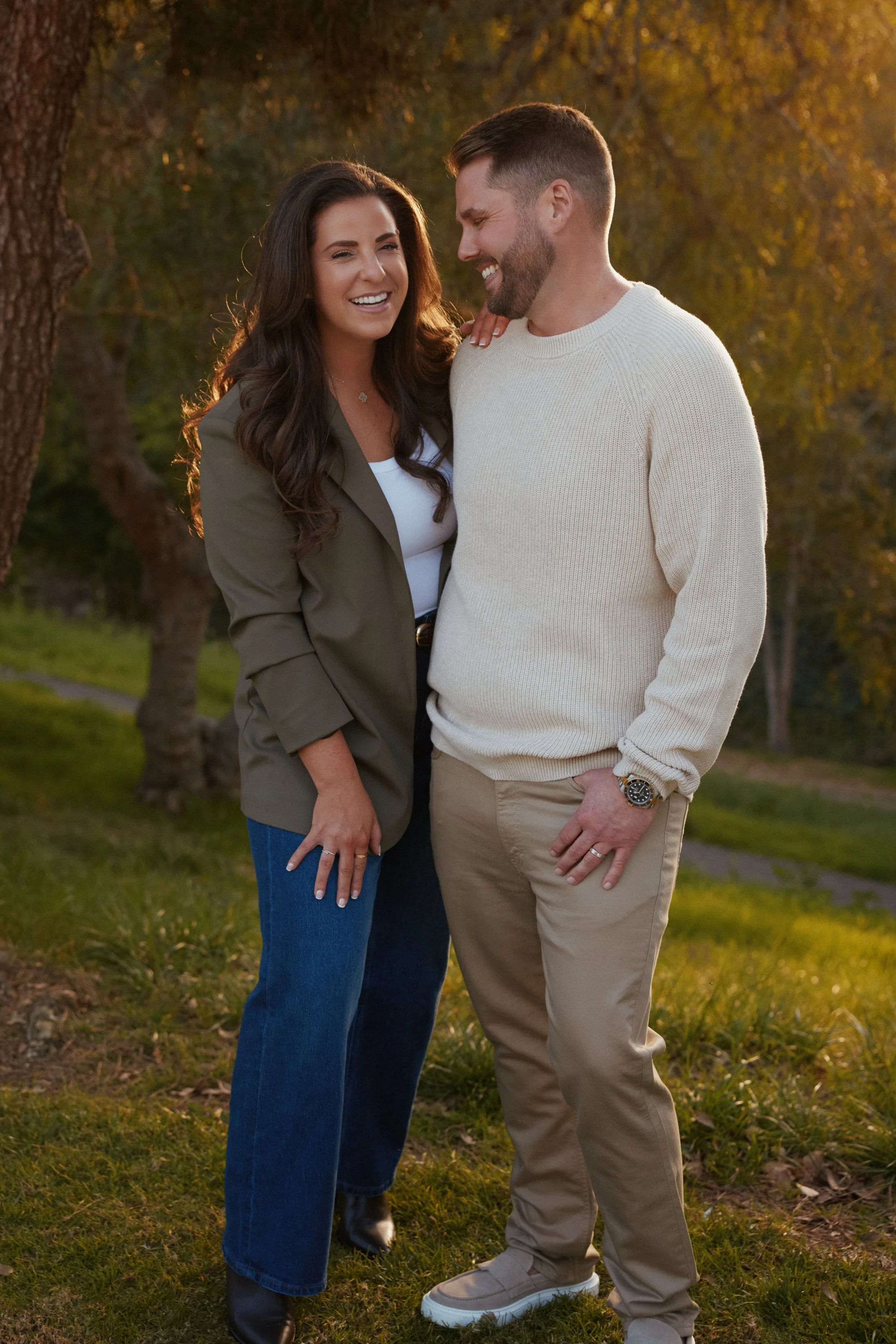 A smiling couple standing outdoors on a grassy area with trees in the background during golden hour. The woman has long brown hair and is wearing a green jacket, white top, and blue jeans. The man has short hair and a beard, wearing a white sweater, tan pants, and a watch. They appear happy and are close to each other.