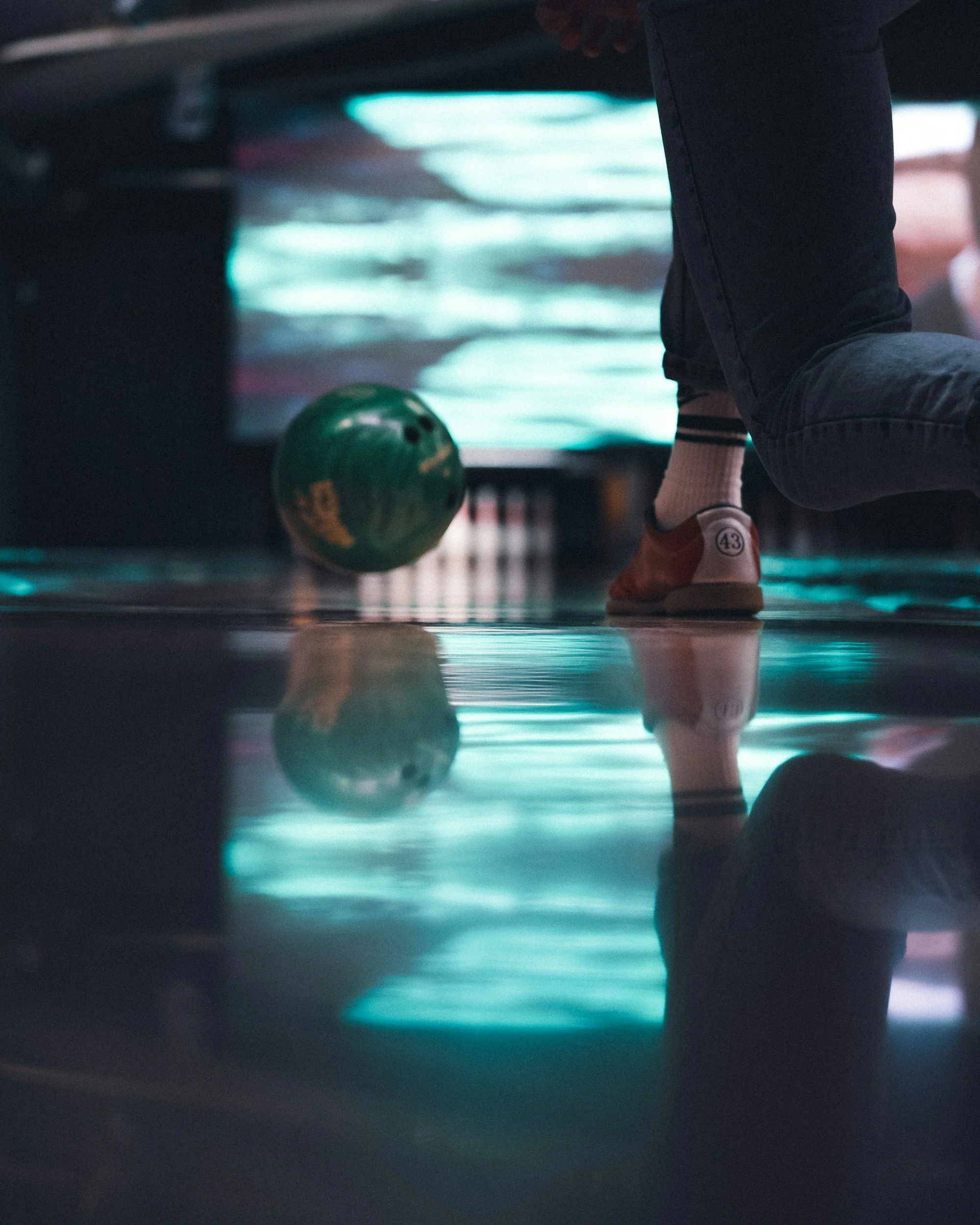 A person about to roll a green bowling ball at a bowling alley, with their reflection visible on the shiny floor.