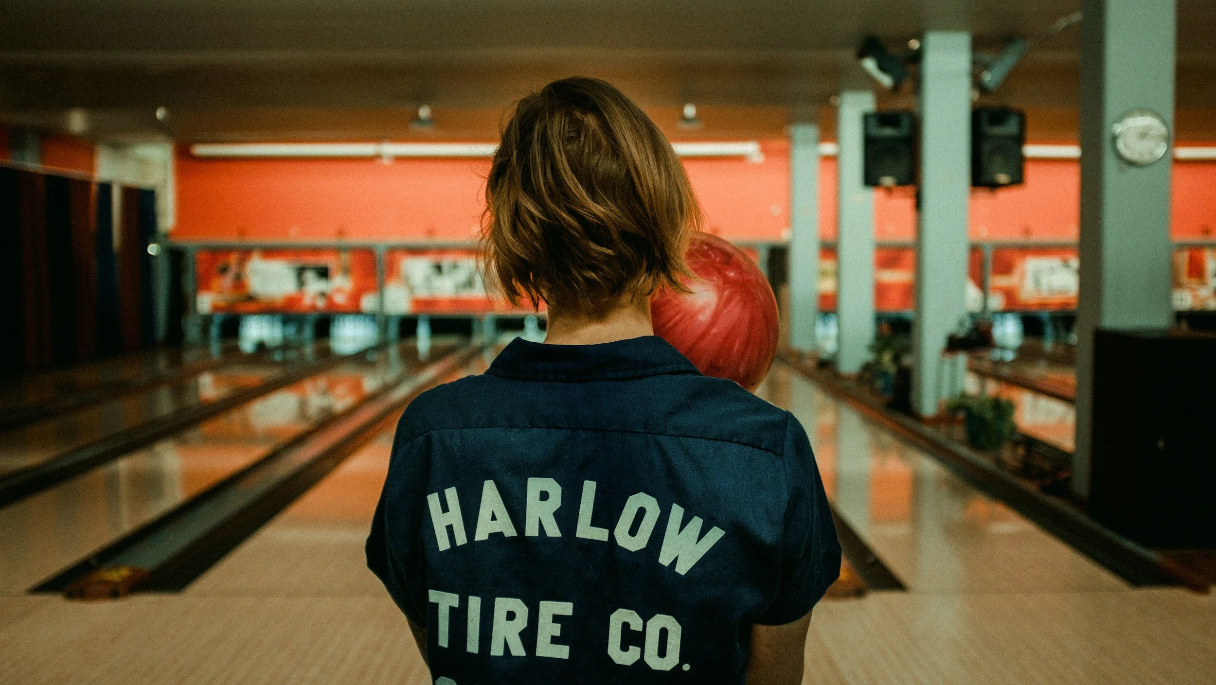 Person with short hair holding a bowling ball in a bowling alley, wearing a jacket with the text "HARLOW TIRE CO." on the back.