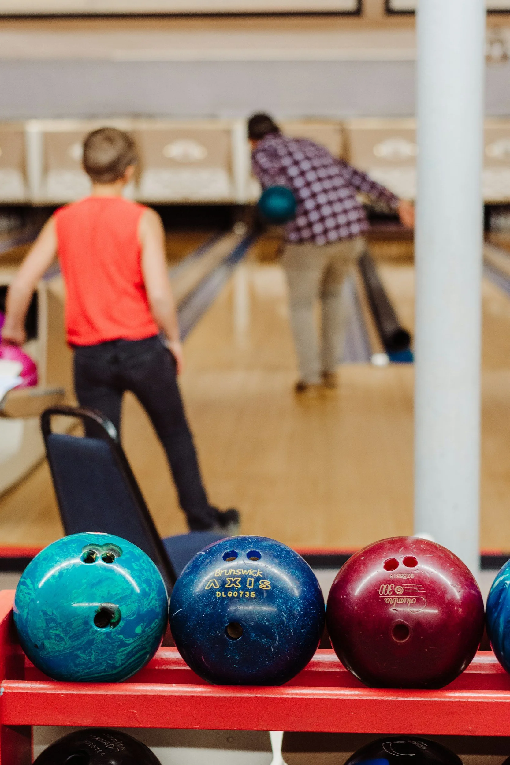 Bowling balls in a red rack with a blurred background of a woman throwing a bowling ball and a young boy watching at a bowling alley.